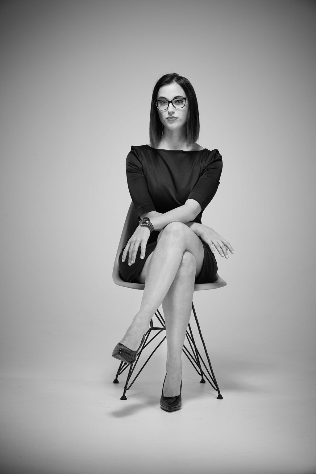 Ein Porträt einer jungen Frau, die auf einem modernen Stuhl sitzt. Sie trägt eine Brille, ein schwarzes Kleid mit Dreiviertelärmeln und hochhackige Schuhe. Ihre Beine sind übereinander geschlagen und sie trägt eine Armbanduhr am linken Handgelenk. Die Frau hat schulterlanges, glattes Haar und schaut direkt in die Kamera mit einem neutralen Gesichtsausdruck. Der Hintergrund ist hell und gleichmäßig beleuchtet.