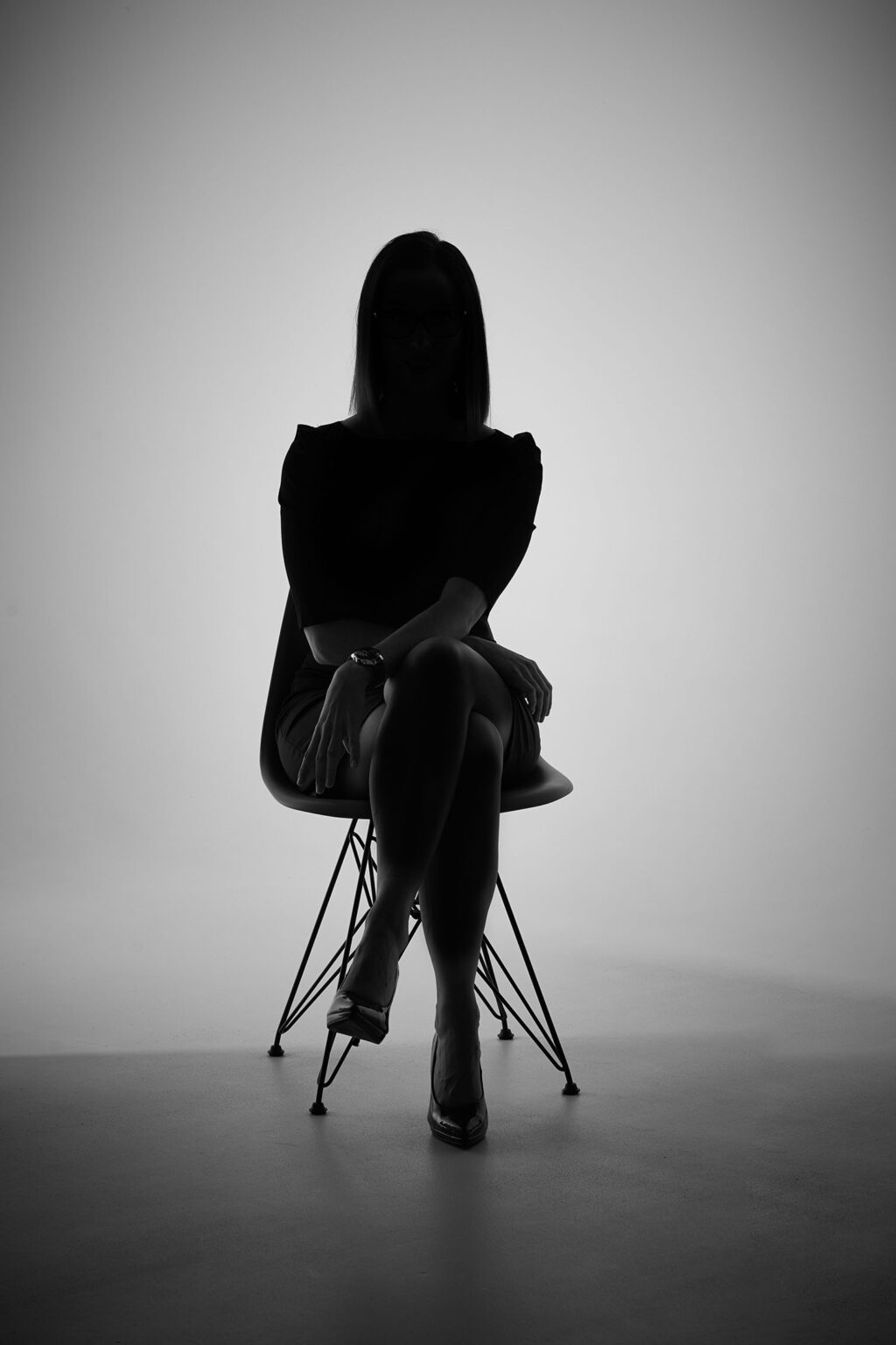 Eine Silhouette einer Frau, die auf einem modernen Stuhl sitzt. Die Frau hat schulterlanges Haar und trägt ein schwarzes Oberteil sowie einen Rock. Ihre Beine sind übereinander geschlagen und sie trägt hochhackige Schuhe. Der Hintergrund ist hell beleuchtet, wodurch die Konturen der Frau stark hervorgehoben werden.