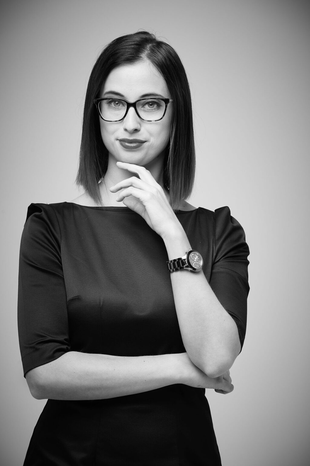 Ein Porträt einer jungen Frau, die direkt in die Kamera schaut. Sie trägt eine Brille, ein schwarzes Kleid mit Dreiviertelärmeln und eine Armbanduhr am linken Handgelenk. Die Frau hat schulterlanges, glattes Haar. Sie hat ihren rechten Arm vor dem Körper verschränkt und berührt mit der rechten Hand ihr Kinn. Der Hintergrund ist hell und gleichmäßig beleuchtet.