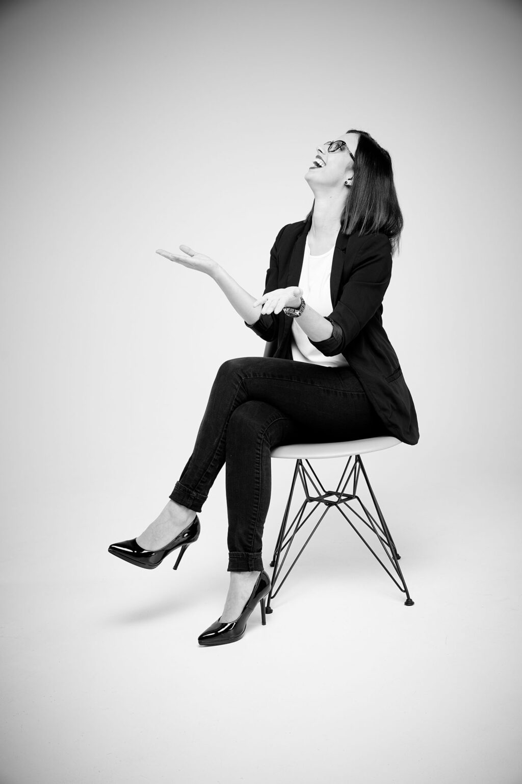 Eine junge Frau sitzt auf einem modernen Stuhl und lacht. Sie trägt eine Brille, ein weißes Oberteil, einen dunklen Blazer, dunkle Jeans und hochhackige Schuhe. Ihre Beine sind übereinandergeschlagen und sie hält die Hände in einer gestikulierenden Bewegung. Der Hintergrund ist hell und gleichmäßig beleuchtet.
