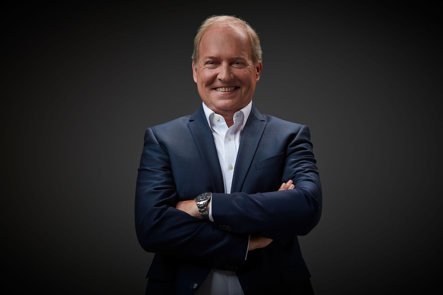 Ein Porträt eines älteren Mannes vor einem dunklen Hintergrund. Er trägt einen dunkelblauen Anzug und ein weißes Hemd. Der Mann hat kurzes, helles Haar und lächelt in die Kamera. Seine Arme sind vor der Brust verschränkt und er trägt eine Armbanduhr am linken Handgelenk.