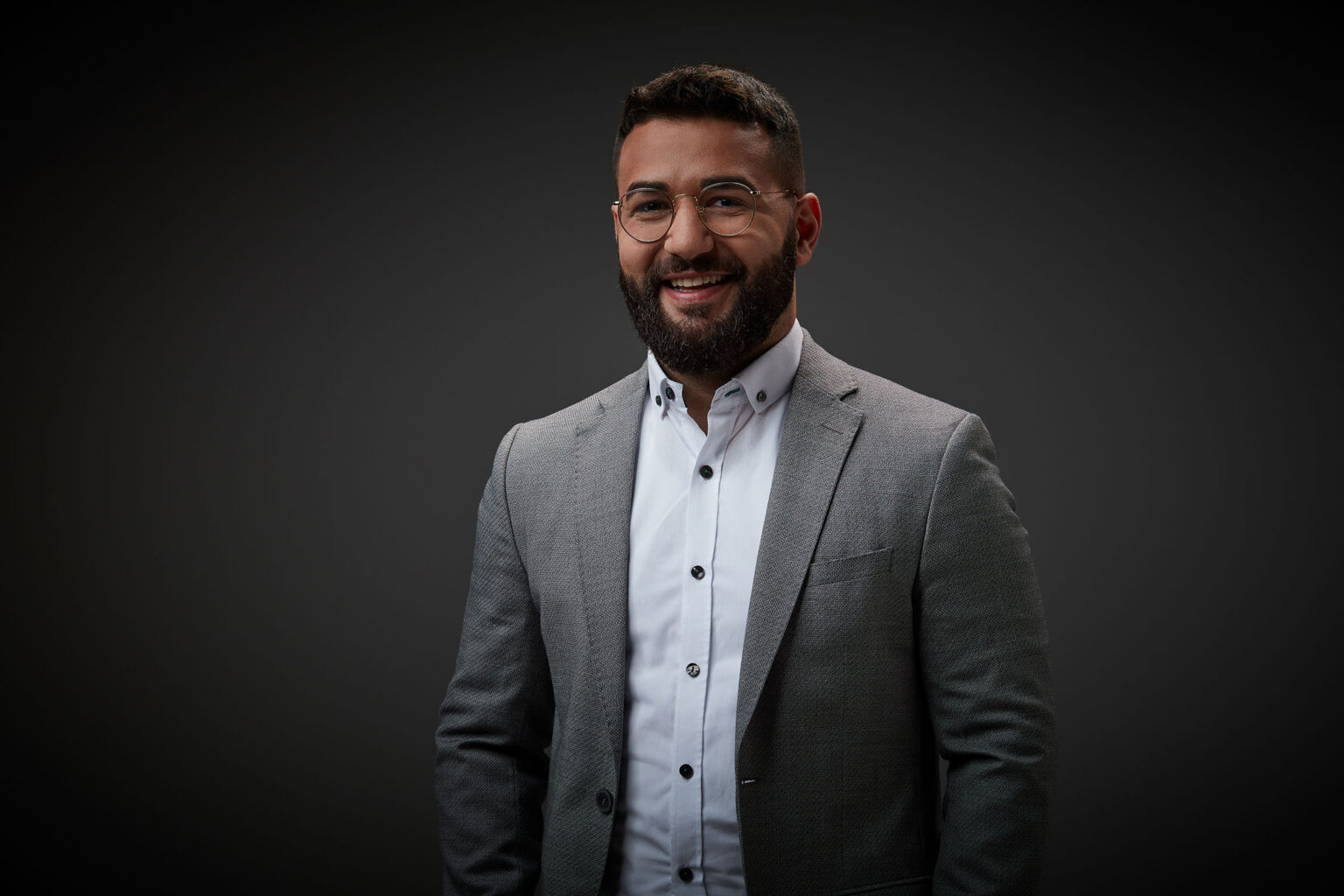 Ein Porträt eines jungen Mannes vor einem dunklen Hintergrund. Er trägt eine Brille, einen grauen Anzug und ein weißes Hemd. Der Mann hat dunkles, kurzes Haar, einen Vollbart und lächelt in die Kamera.