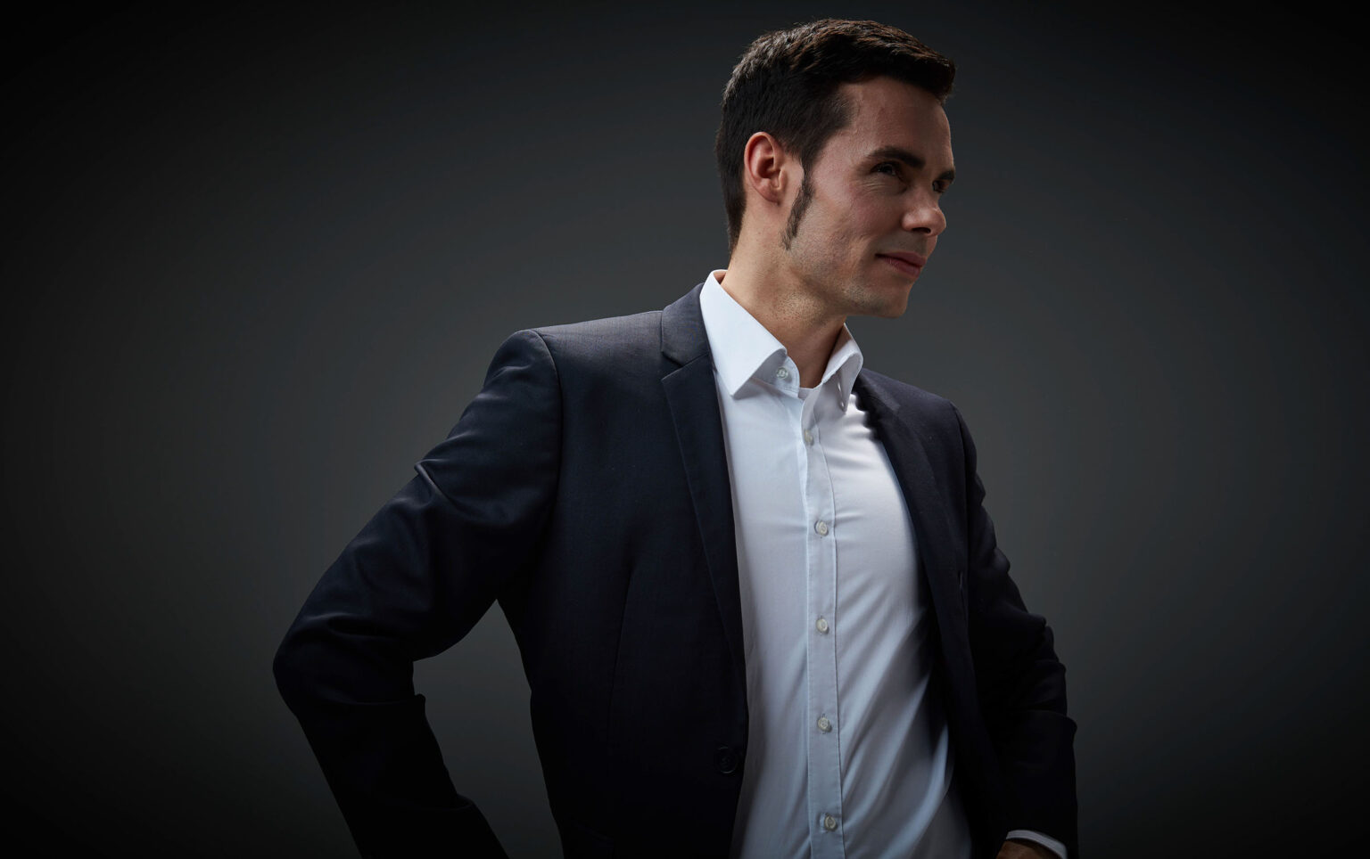 Ein Porträt eines jungen Mannes in seitlicher Ansicht vor einem dunklen Hintergrund. Er trägt einen dunkelblauen Anzug und ein weißes Hemd. Der Mann hat dunkles, kurzes Haar und schaut leicht lächelnd zur Seite. Eine Hand ist in die Hüfte gestützt.