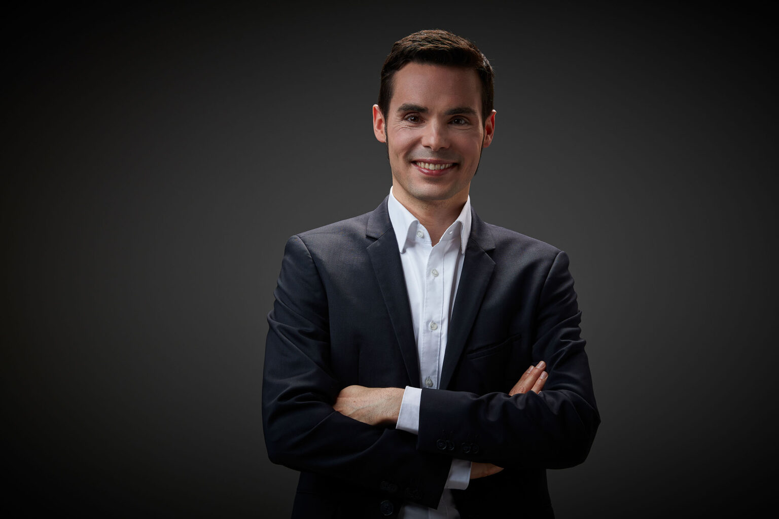 Ein Porträt eines jungen Mannes vor einem dunklen Hintergrund. Er trägt einen dunkelblauen Anzug und ein weißes Hemd. Der Mann hat dunkles, kurzes Haar und lächelt in die Kamera. Seine Arme sind vor der Brust verschränkt.