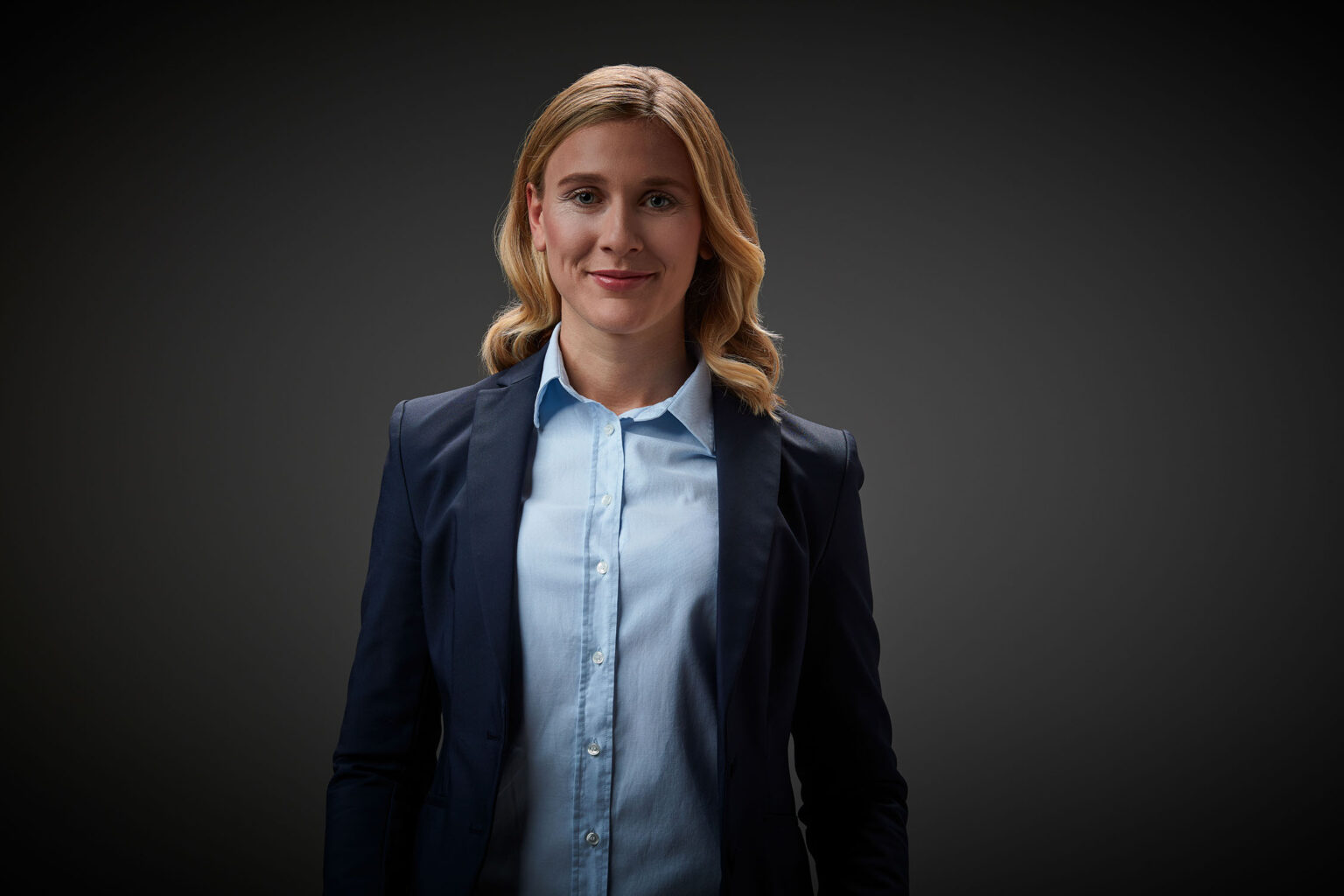 Ein Porträt einer jungen Frau vor einem dunklen Hintergrund. Sie trägt einen dunkelblauen Blazer und ein hellblaues Hemd. Die Frau hat schulterlanges, blondes, gewelltes Haar und lächelt leicht in die Kamera.