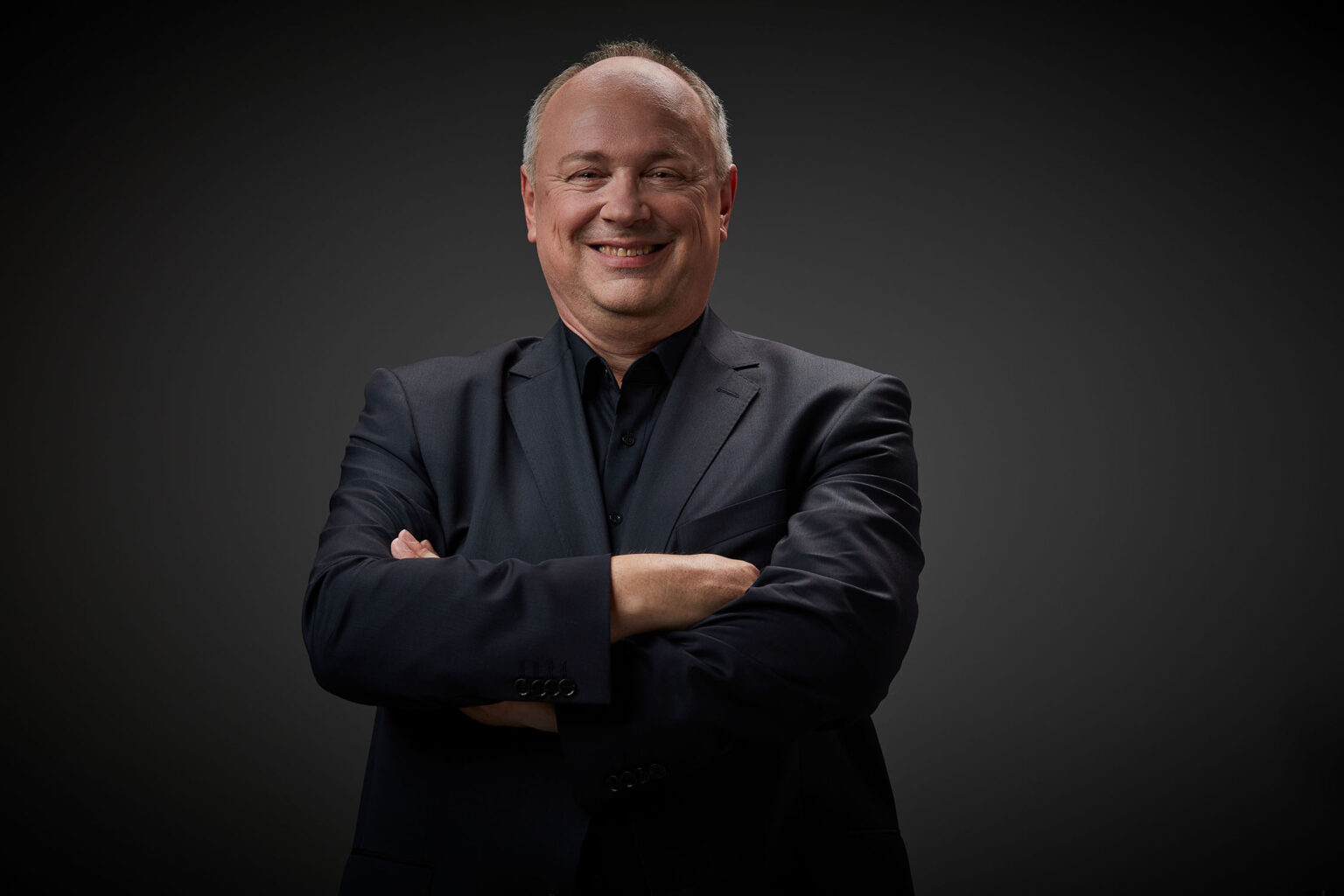 Ein Porträt eines älteren Mannes vor einem dunklen Hintergrund. Er trägt einen dunkelblauen Anzug und ein schwarzes Hemd. Der Mann hat kurzes, helles Haar und lächelt in die Kamera. Seine Arme sind vor der Brust verschränkt.