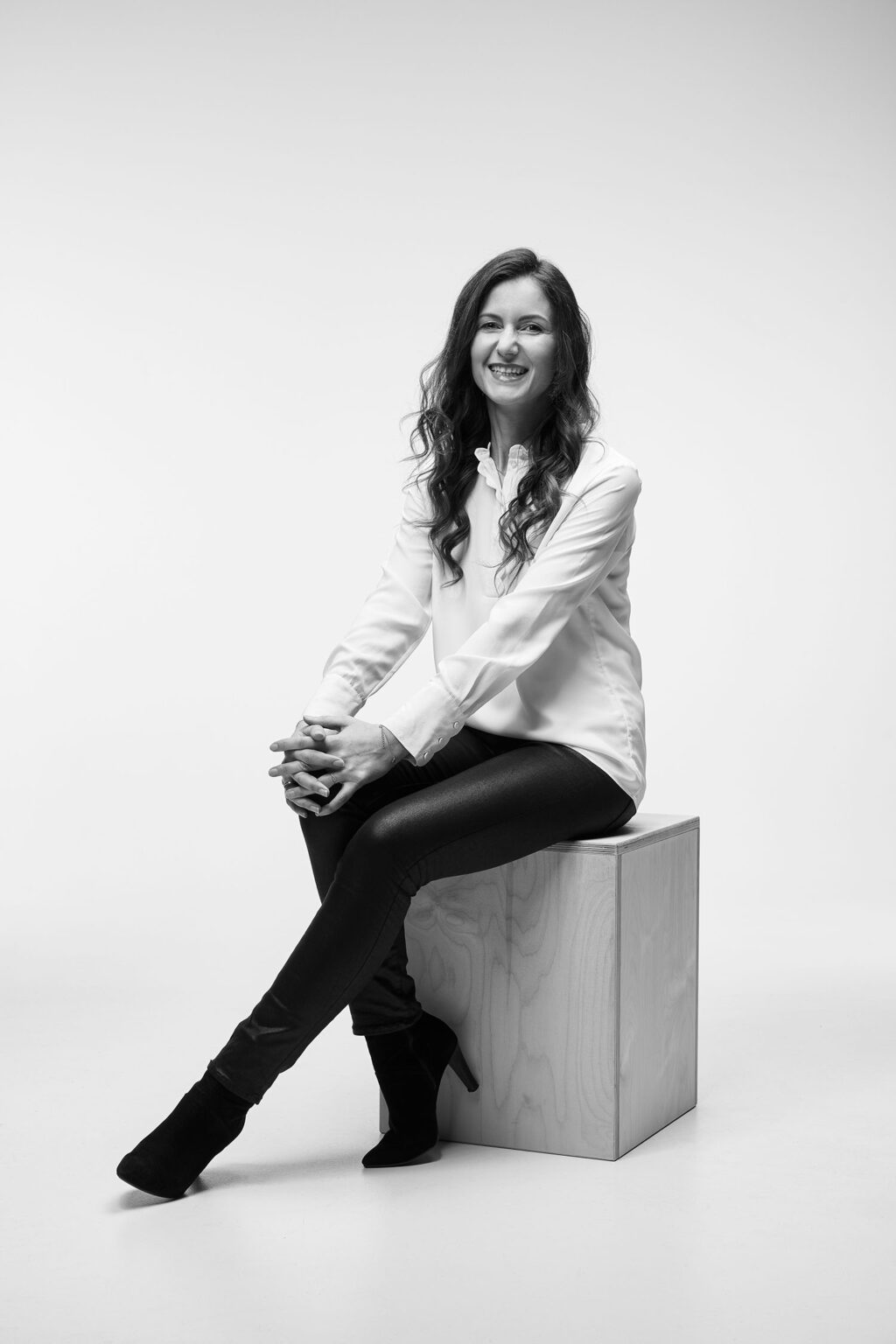 Eine junge Frau mit langen, lockigen Haaren sitzt auf einem Holzwürfel vor einem weißen Hintergrund. Sie trägt eine helle Bluse, dunkle Hosen und dunkle Stiefeletten mit Absätzen. Ihre Beine sind übereinander geschlagen und ihre Hände ruhen auf ihrem Knie. Die Frau lächelt in die Kamera. Das Bild ist in Schwarz-Weiß aufgenommen.