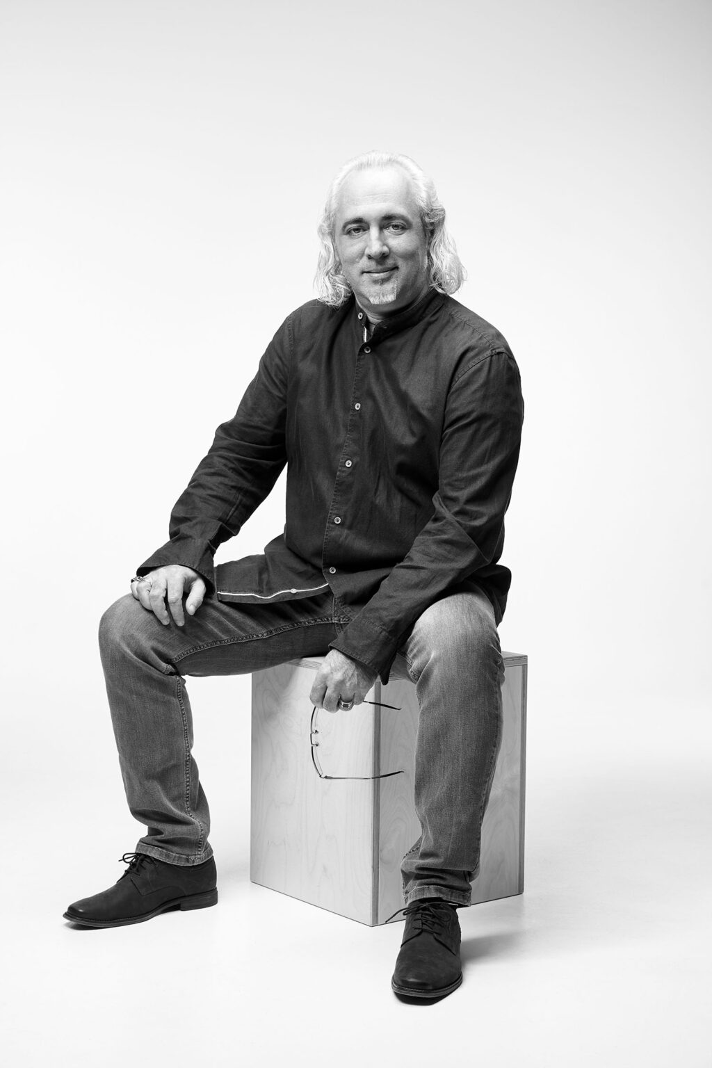 Ein Mann mittleren Alters mit schulterlangen grauen Haaren sitzt auf einem Holzwürfel vor einem weißen Hintergrund. Er trägt ein dunkles Hemd, Jeans und dunkle Schuhe. Der Mann hält eine Brille in seiner rechten Hand und lächelt leicht in die Kamera. Das Bild ist in Schwarz-Weiß aufgenommen.