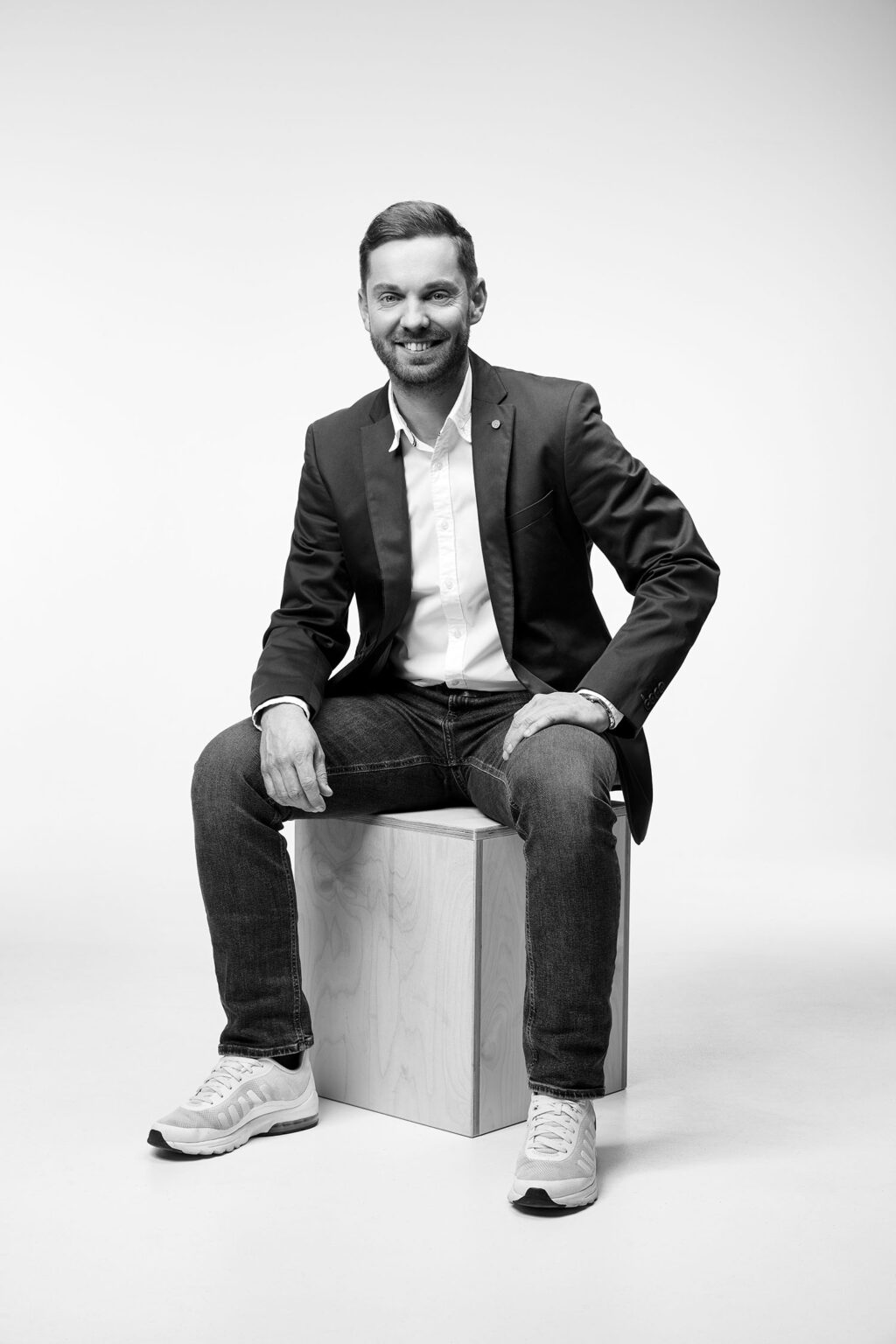 Ein junger Mann sitzt auf einem Holzwürfel vor einem weißen Hintergrund. Er trägt ein dunkles Sakko, ein weißes Hemd, Jeans und helle Sportschuhe. Der Mann lächelt in die Kamera und hat eine entspannte Haltung. Das Bild ist in Schwarz-Weiß aufgenommen.