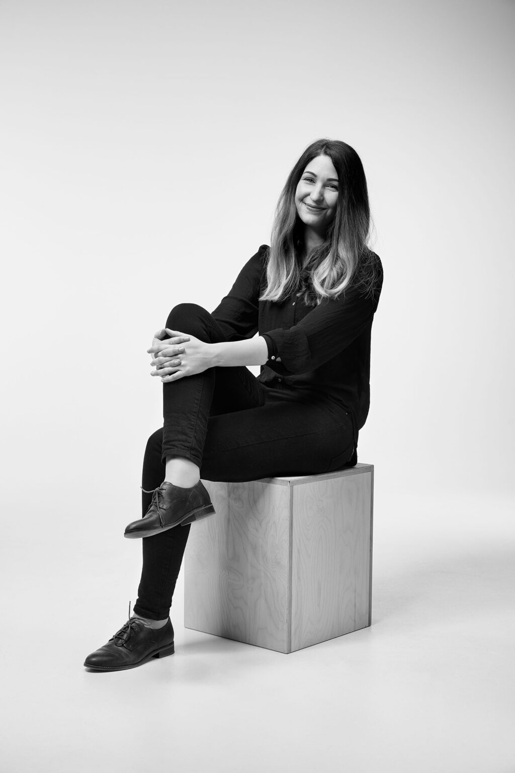 Eine junge Frau mit langen Haaren sitzt auf einem Holzwürfel vor einem weißen Hintergrund. Sie trägt eine dunkle Bluse, dunkle Hosen und dunkle Schuhe. Ihre Beine sind übereinander geschlagen und ihre Hände ruhen auf ihrem Knie. Die Frau lächelt leicht in die Kamera. Das Bild ist in Schwarz-Weiß aufgenommen.