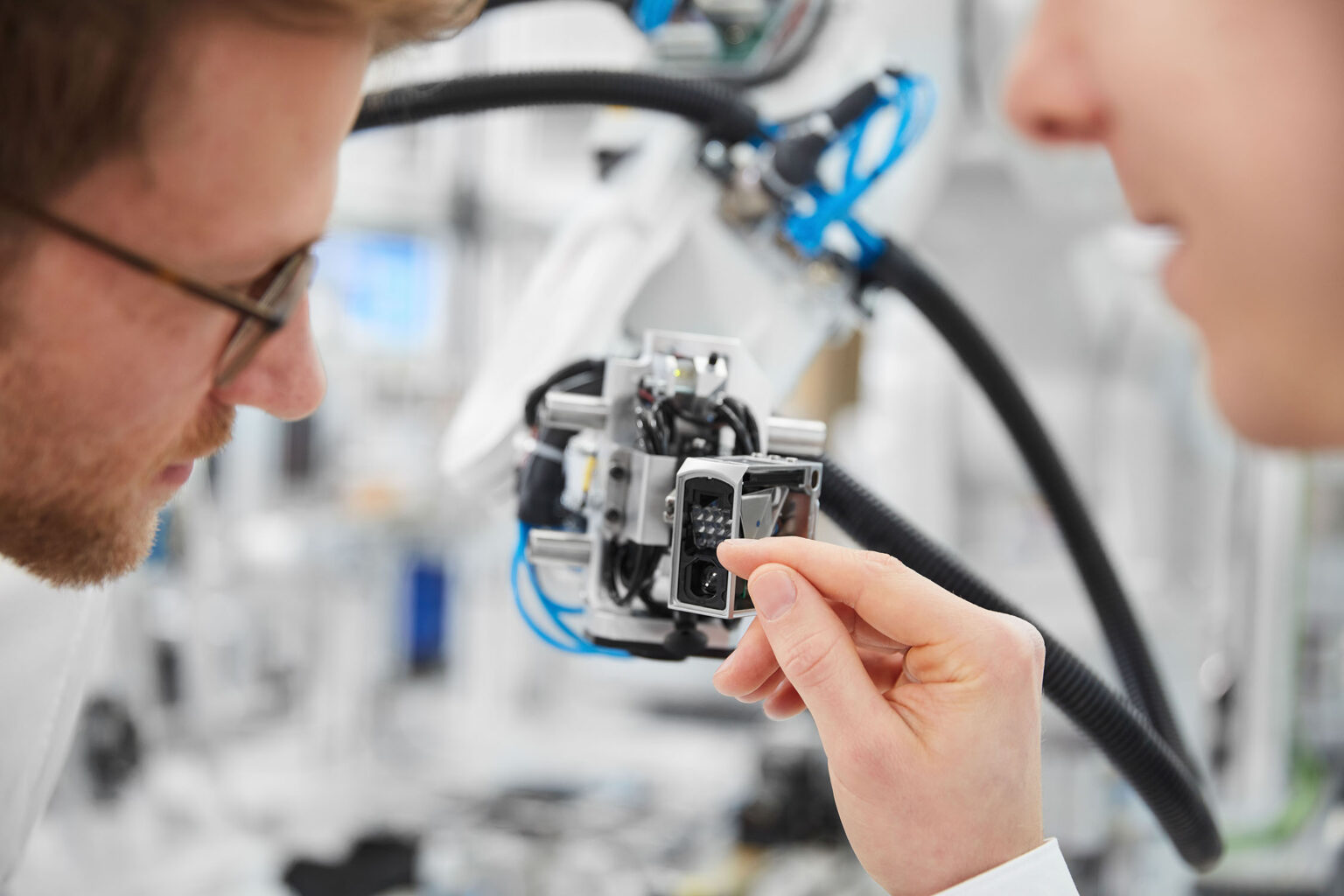 Zwei Personen arbeiten an einer Maschine in einer modernen Produktionsumgebung. Die Person links, teilweise unscharf im Vordergrund, trägt eine Brille und schaut auf ein kleines Bauteil, das die Person rechts, ebenfalls unscharf im Vordergrund, in der Hand hält. Im Hintergrund sind verschiedene Maschinen und technische Ausrüstungen mit blauen und schwarzen Kabeln zu sehen.