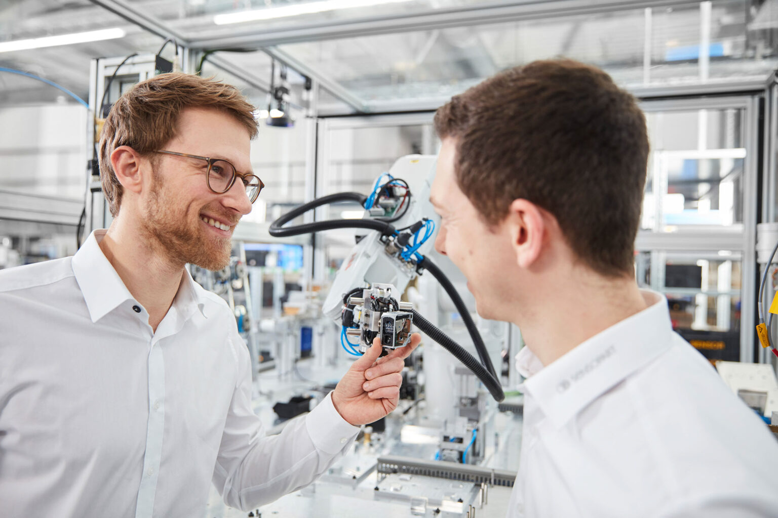 Zwei Männer in weißen Hemden stehen in einer modernen Produktionsumgebung und lächeln einander an. Der Mann links trägt eine Brille und hält ein kleines technisches Bauteil in der Hand. Der Mann rechts, der ebenfalls lächelt, schaut auf das Bauteil. Im Hintergrund sind Maschinen und technische Ausrüstungen mit Kabeln und Schläuchen zu sehen.