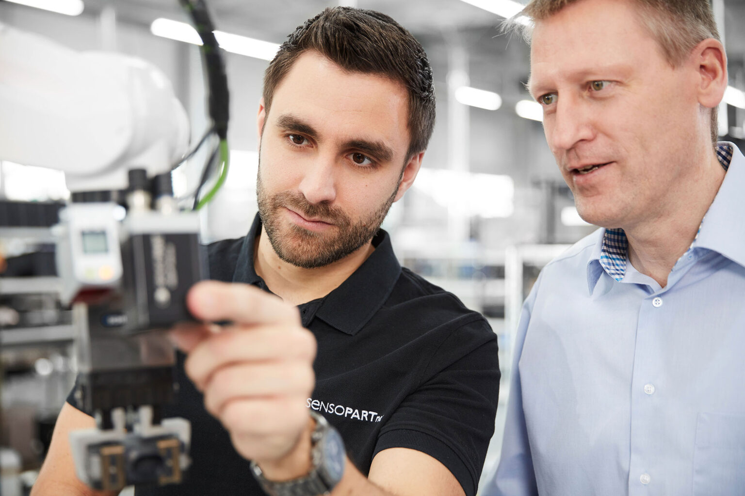 Zwei Männer arbeiten gemeinsam in einer modernen Produktionsumgebung. Der Mann links, der einen dunklen Bart und ein schwarzes Polohemd mit der Aufschrift 'SensoPart' trägt, hält ein technisches Gerät und schaut konzentriert darauf. Der Mann rechts trägt ein hellblaues Hemd und schaut ebenfalls auf das Gerät. Im Hintergrund sind unscharfe Details der Produktionshalle zu sehen.