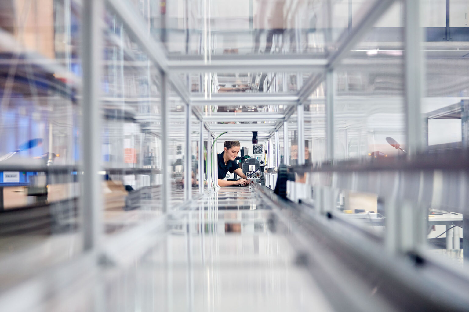 Eine Frau in Arbeitskleidung arbeitet an einer Maschine in einer modernen Produktionsanlage. Die Perspektive des Bildes zeigt eine lange, lineare Anordnung von Metallrahmen, die die Tiefe der Anlage betonen. Die Frau ist konzentriert bei der Arbeit und blickt auf die Maschine vor ihr. Im Hintergrund sind unscharfe Details weiterer Maschinen und Produktionsausrüstung zu sehen.