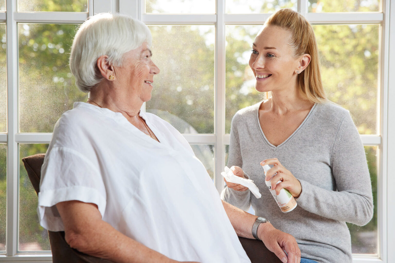 Eine ältere Frau mit weißem Haar und einem weißen Oberteil sitzt und lächelt eine jüngere Frau mit zurückgebundenem Haar an, die in einem grauen Pullover vor einem Fenster steht. Die jüngere Frau hält eine Flasche und ein Tuch in den Händen und scheint im Begriff zu sein, etwas zu reinigen oder zu pflegen. Das Zimmer ist hell, mit Tageslicht, das durch das Fenster fällt, was eine warme und behagliche Atmosphäre schafft.