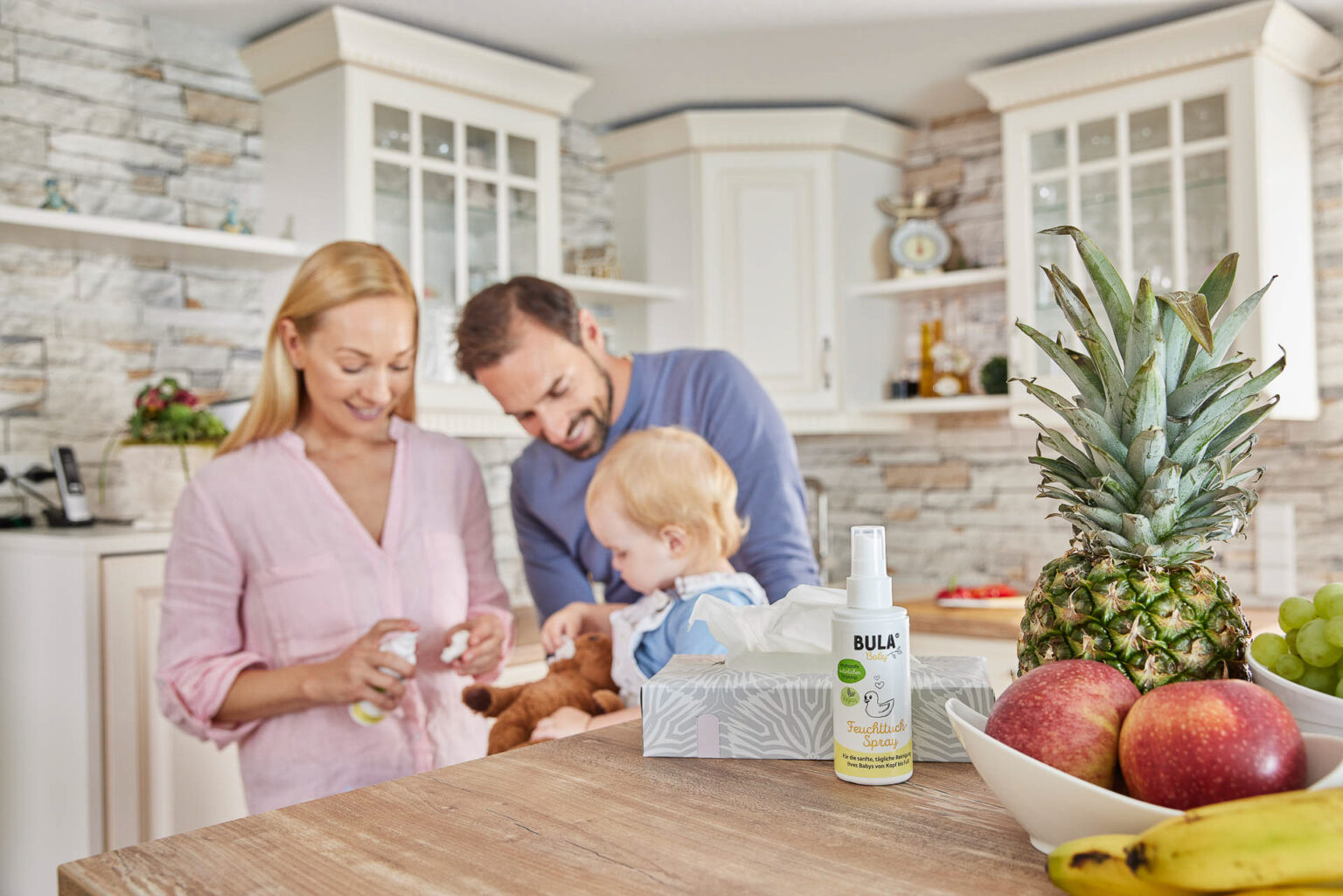 Eine Familie genießt gemeinsam eine Aktivität in einer gemütlichen Küche. Eine blonde Frau in einem rosa Hemd gibt einem kleinen Mädchen, das auf dem Küchentresen sitzt, ein Tuch, während ein Mann in einem blauen Shirt liebevoll zusehen. Auf dem Tresen stehen eine Flasche Feuchtigkeitsspray und eine Box mit Tüchern neben einer Schale mit frischem Obst und einer Ananas. Die Szene strahlt Wärme, Fürsorge und häusliches Glück aus.