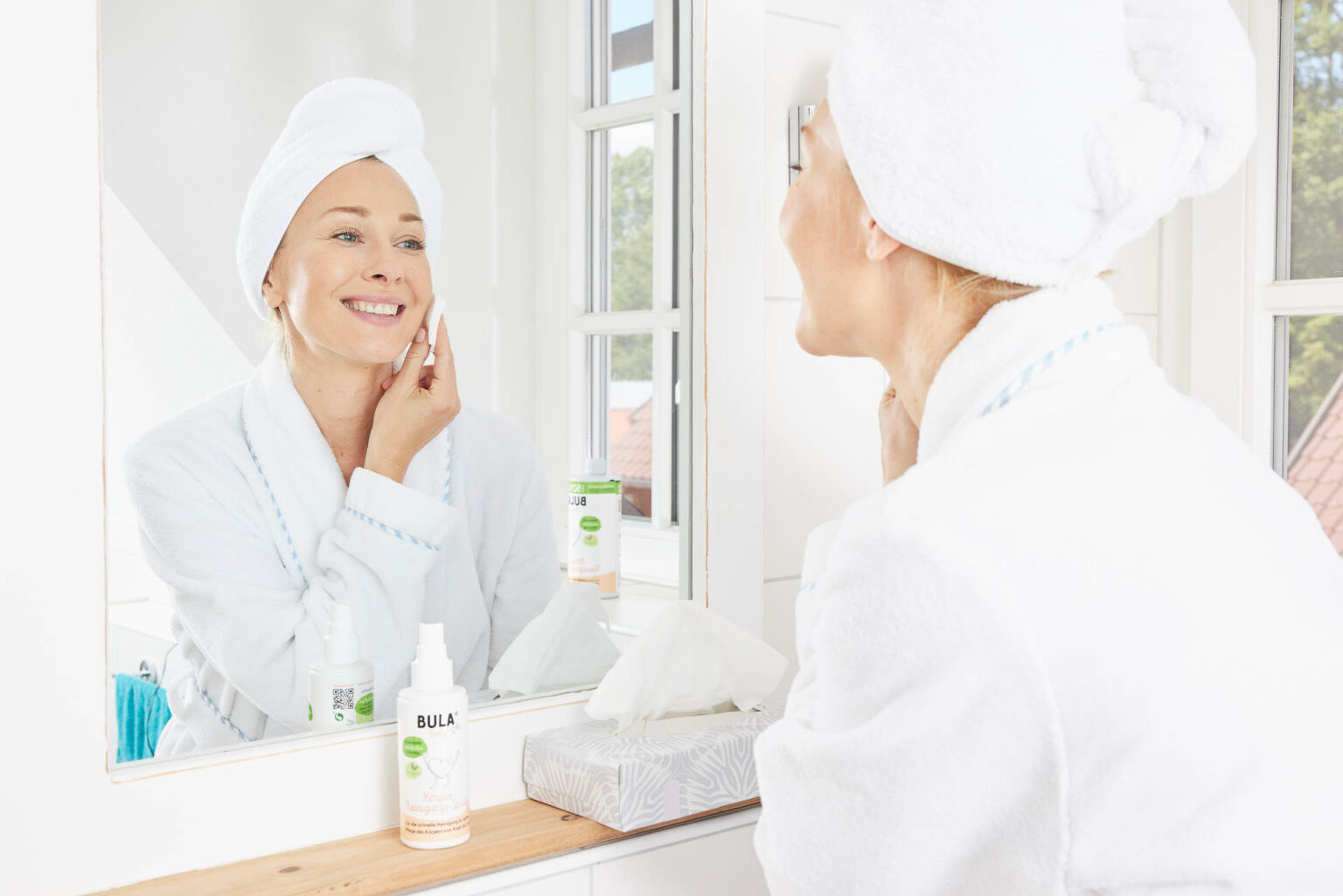 Eine Frau in einem weißen Bademantel und mit einem Handtuchturban auf dem Kopf steht in einem hellen Badezimmer und lächelt ihr Spiegelbild an, während sie eine Gesichtspflegeflasche in einer Hand hält und sanft mit der anderen Hand ihre Wange berührt. Auf der Arbeitsfläche vor ihr befinden sich eine weitere Pflegeflasche und eine Box mit Kosmetiktüchern. Das Bild vermittelt ein Gefühl von Frische und Pflege in einem privaten, wohnlichen Umfeld.