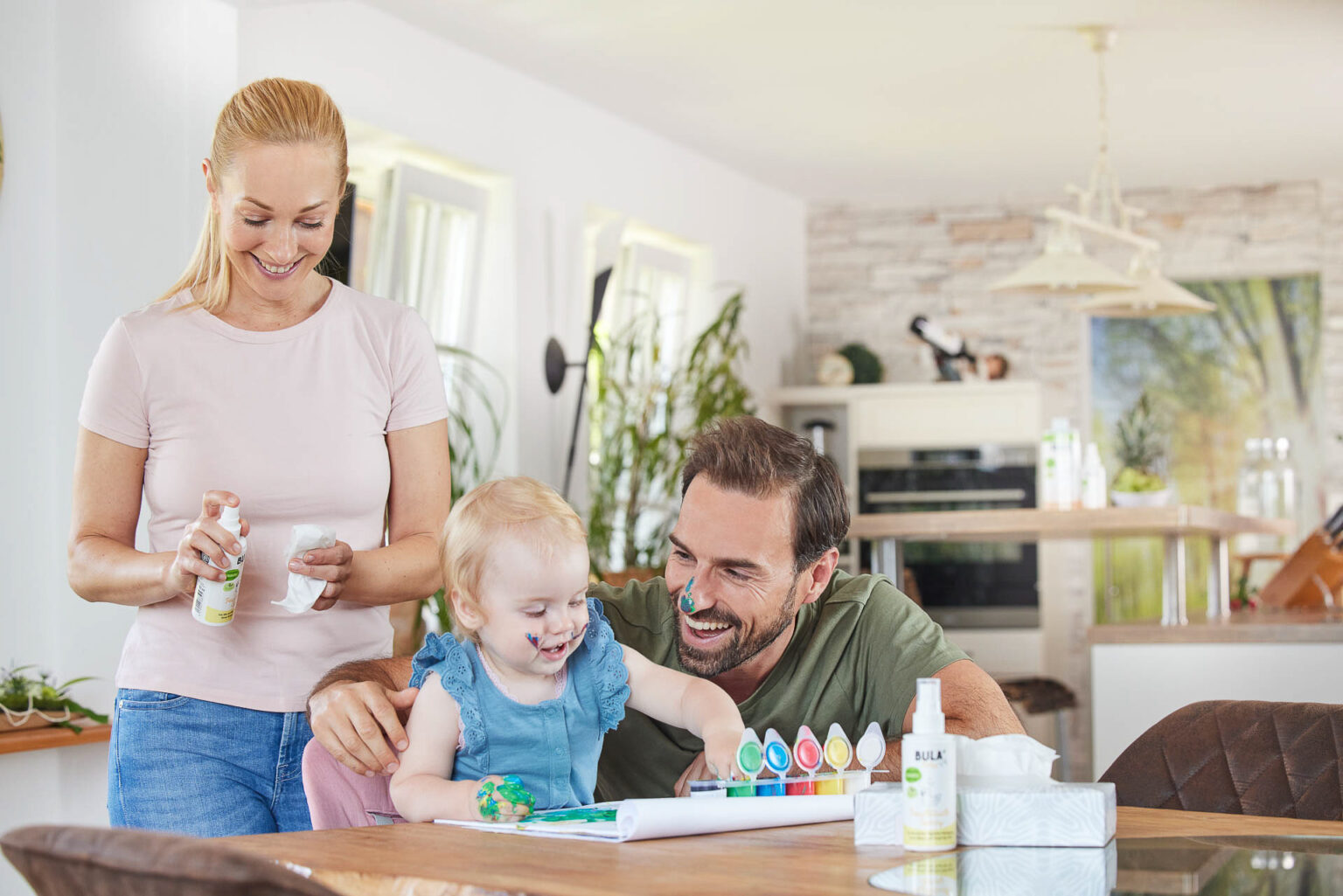 Ein helles und freundliches Familienbild in einem gemütlichen Innenraum. Eine blonde Frau im rosa T-Shirt hält eine Sprühflasche und ein Tuch, während sie sich über ein kleines Mädchen mit blonden Haaren beugt, das auf einem Stuhl sitzt und begeistert Farben auswählt. Neben ihnen steht ein lachender Mann im grünen Shirt, der dem Mädchen hilft, während Farbtuben und Bastelmaterialien auf dem Holztisch verteilt sind. Die Szene strahlt Wärme und familiäre Geborgenheit aus.