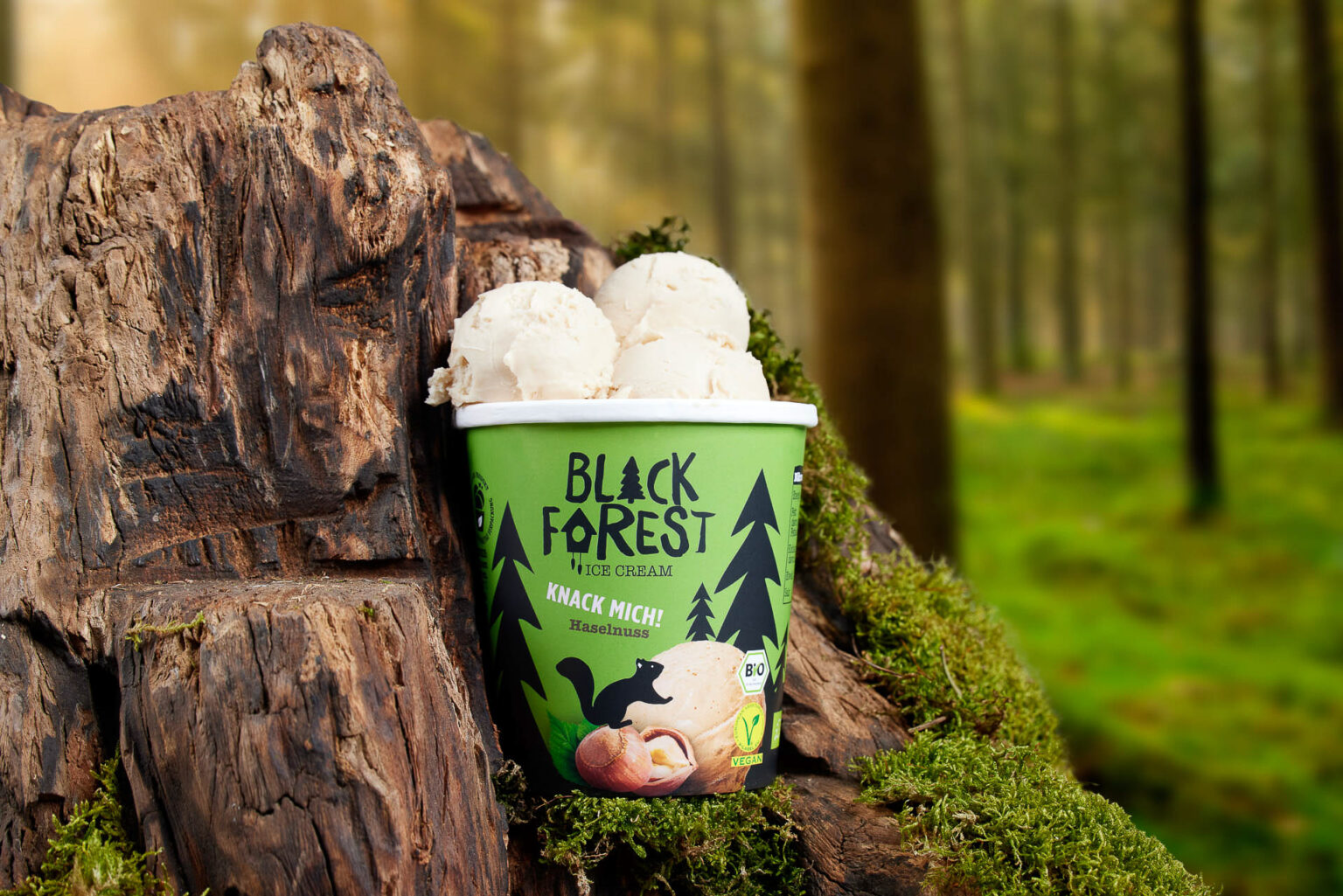 Ein Becher mit Haselnuss-Eiskrem steht auf einem moosbedeckten Baumstamm in einem lichten Wald. Der Becher ist lebhaft grün mit dem Logo "BLACK FOREST ICE CREAM" und der Aufschrift "KNACK MICH! Haselnuss", zudem sind Bio- und Vegan-Siegel sichtbar. Die Kugeln Eiskrem sehen cremig und einladend aus, die Umgebung ist natürlich und friedlich mit weichem Sonnenlicht, das durch die Bäume scheint.