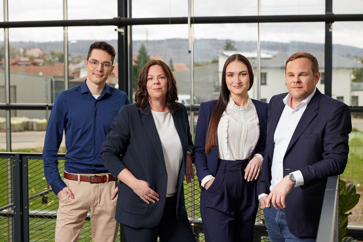 Eine Gruppe von vier Personen steht vor einer großen Fensterfront in einem modernen Bürogebäude. Von links nach rechts: Ein junger Mann mit Brille, der ein blaues Hemd und beige Hosen trägt, eine Frau mit schulterlangem braunen Haar in einem dunklen Blazer und weißem Oberteil, eine junge Frau mit langen braunen Haaren in einer weißen Bluse mit Rüschen und einem dunkelblauen Blazer, und ein Mann mit kurzen blonden Haaren in einem weißen Hemd und dunklem Blazer. Im Hintergrund sind Gebäude und eine Landschaft zu sehen.