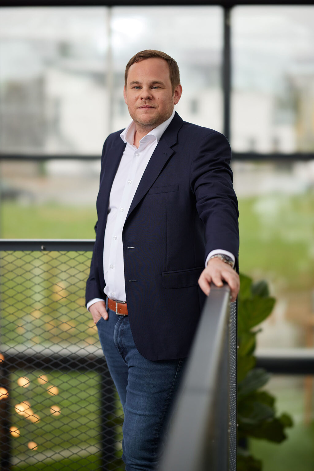 Ein Mann mit kurzen blonden Haaren steht in einem modernen Bürogebäude. Er trägt ein weißes Hemd, einen dunklen Blazer und eine Jeans. Der Mann lehnt sich an ein Geländer und schaut in die Kamera. Im Hintergrund sind unscharf Fenster und eine Landschaft zu sehen.