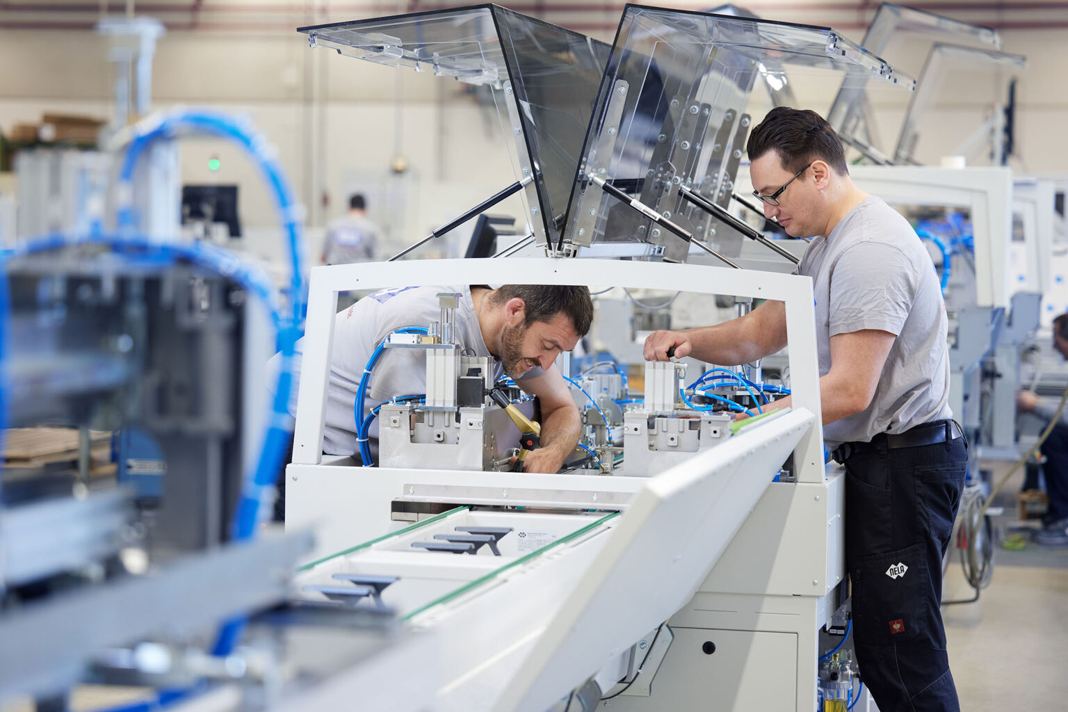 Zwei Männer in grauen T-Shirts arbeiten an einer großen Maschine in einer industriellen Produktionsumgebung. Der Mann links beugt sich über die Maschine und führt eine manuelle Tätigkeit aus, während der Mann rechts, der eine Brille trägt, ebenfalls an der Maschine arbeitet und ein Bauteil justiert. Die Maschine ist mit blauen Kabeln ausgestattet und hat geöffnete transparente Abdeckungen. Im Hintergrund sind weitere Maschinen und Produktionsausrüstung zu sehen.