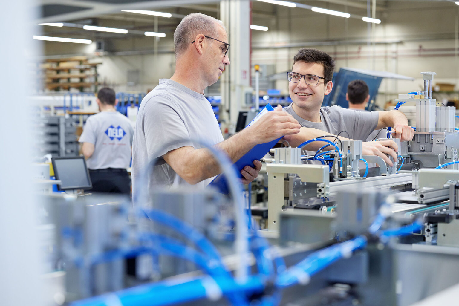 Zwei Männer in grauen T-Shirts arbeiten in einer industriellen Produktionsumgebung. Der Mann links, der eine Brille trägt, hält ein blaues Klemmbrett und spricht mit dem Mann rechts, der ebenfalls eine Brille trägt und lächelt. Beide Männer stehen vor einer Maschine mit blauen Kabeln. Im Hintergrund sind weitere Personen und industrielle Ausrüstungen zu sehen.