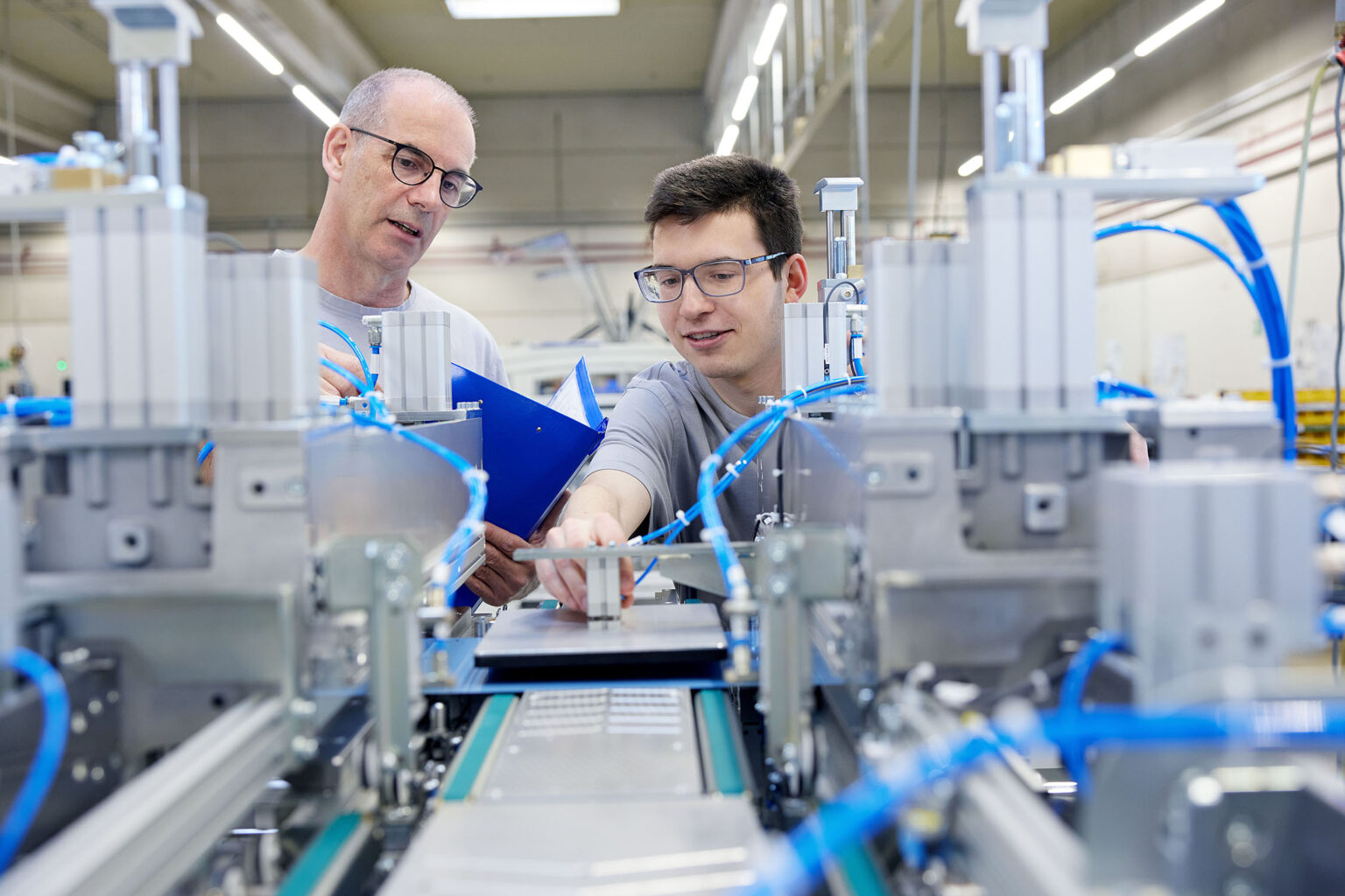 Zwei Männer in grauen T-Shirts arbeiten gemeinsam an einer Maschine in einer industriellen Produktionsumgebung. Der Mann links trägt eine Brille und hält ein blaues Klemmbrett, während er mit dem anderen Mann spricht. Der Mann rechts, der ebenfalls eine Brille trägt, zeigt auf ein Bauteil der Maschine. Die Maschine ist mit blauen Kabeln ausgestattet, und im Hintergrund sind weitere industrielle Ausrüstungen zu sehen.