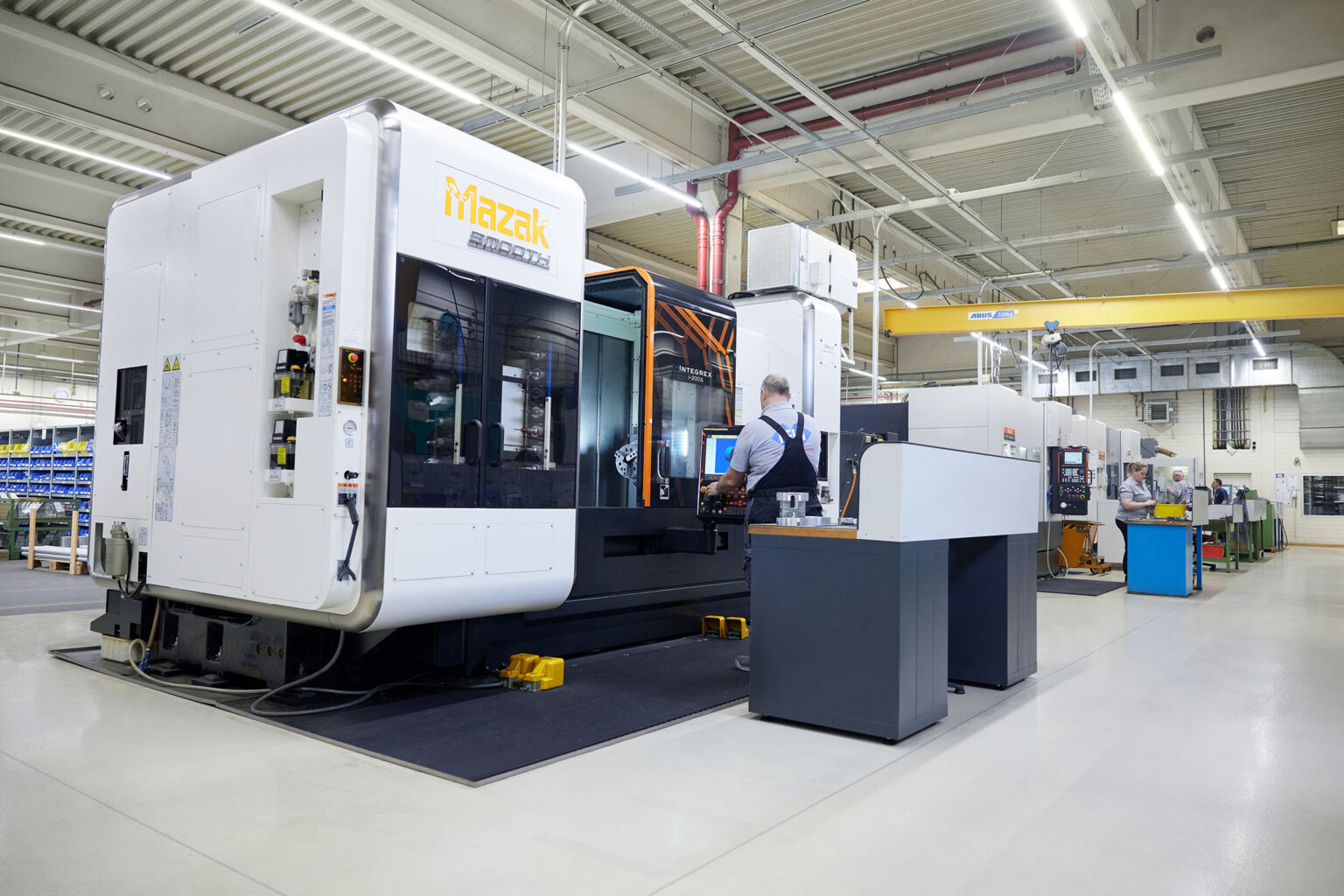 Eine industrielle Produktionshalle mit mehreren großen Maschinen. Im Vordergrund befindet sich eine große, weiße Maschine mit der Aufschrift 'Mazak' und ein Bediener in Arbeitskleidung, der an einem Kontrollpanel arbeitet. Im Hintergrund sind weitere Maschinen, Arbeitsstationen und Arbeiter in der Halle zu sehen. Die Halle ist gut beleuchtet und hat eine strukturierte Decke mit sichtbaren Leitungen und Beleuchtung.