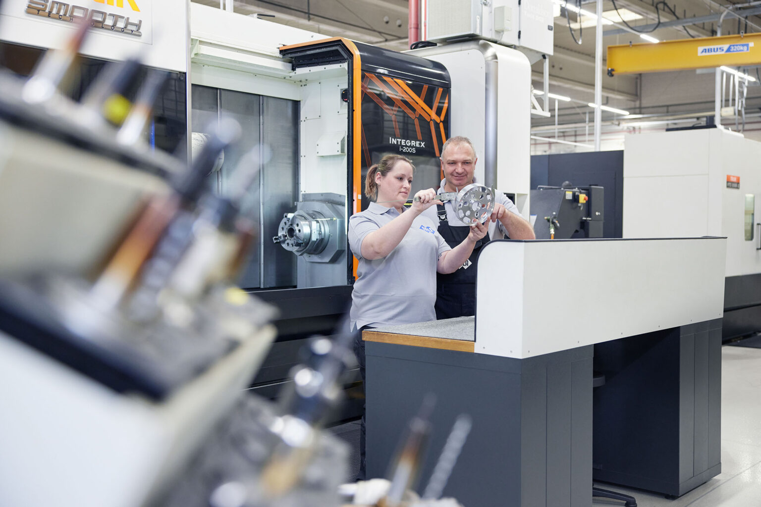 Zwei Personen in grauen Arbeitskleidung stehen vor einer großen Maschine in einer industriellen Produktionsumgebung. Die Frau links hält ein metallisches Bauteil und ein Messwerkzeug, während der Mann rechts ihr zeigt, wie es verwendet wird. Beide konzentrieren sich auf das Bauteil. Im Hintergrund ist eine Maschine mit der Aufschrift 'Integrex' und 'Mazak' sowie weitere Produktionsausrüstung zu sehen.