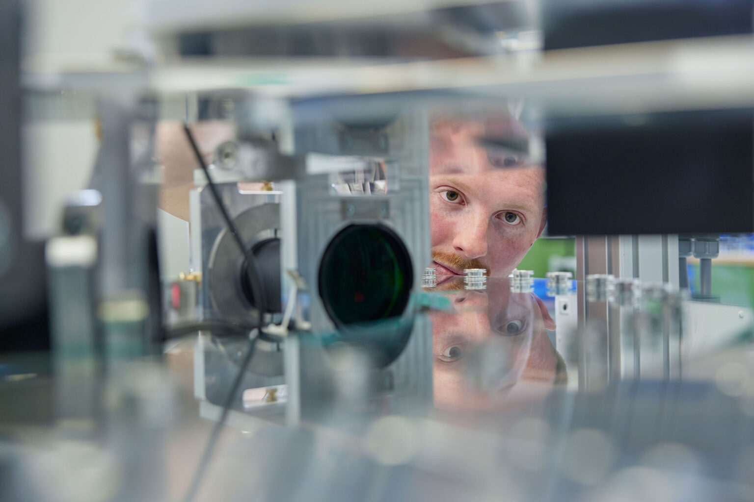 Ein Mann schaut konzentriert auf eine Maschine in einer industriellen Produktionsumgebung. Sein Gesicht spiegelt sich in den glänzenden Oberflächen der Maschine. Die Maschine ist mit verschiedenen technischen Komponenten und Kabeln ausgestattet. Der Fokus liegt auf dem Gesicht des Mannes und den präzisen Details der Maschine.