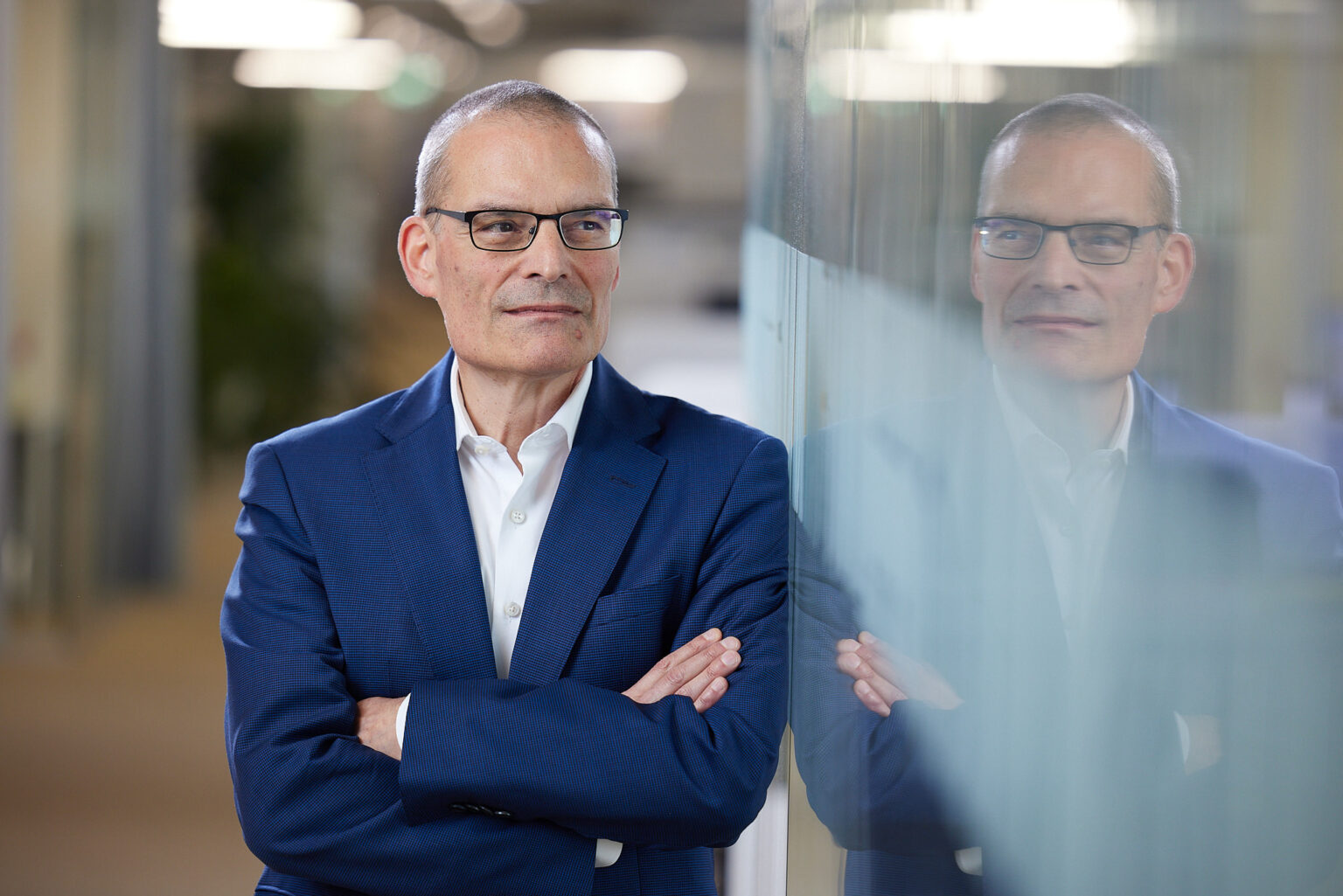 Ein selbstbewusster, älterer Herr mit grauem Haar und Brille steht mit verschränkten Armen und einem leichten Lächeln im Gesicht. Er trägt ein elegantes dunkelblaues Sakko über einem weißen Hemd. Sein Spiegelbild ist in einer Glaswand sichtbar, die Teil eines modernen Büroinnenraums zu sein scheint. Das Licht im Raum schafft einen warmen, einladenden Effekt.