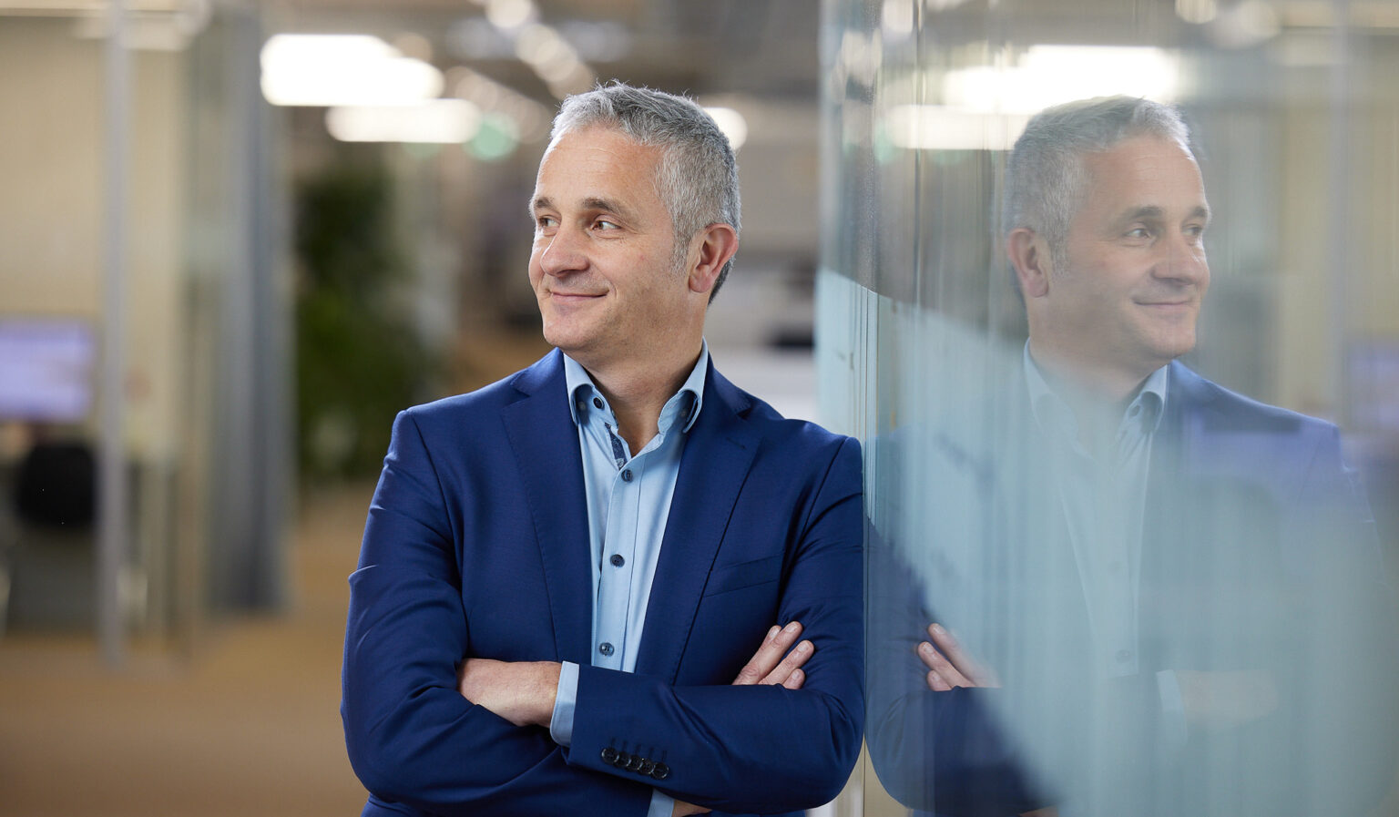 Ein Mann mittleren Alters mit grauen Haaren steht mit verschränkten Armen und lächelt. Er trägt ein blaues Sakko und ein hellblaues Hemd. Der Mann lehnt an einer Glaswand, die sein Spiegelbild zeigt. Im Hintergrund ist ein modernes Büro mit unscharfen Lichtquellen und Pflanzen zu sehen.