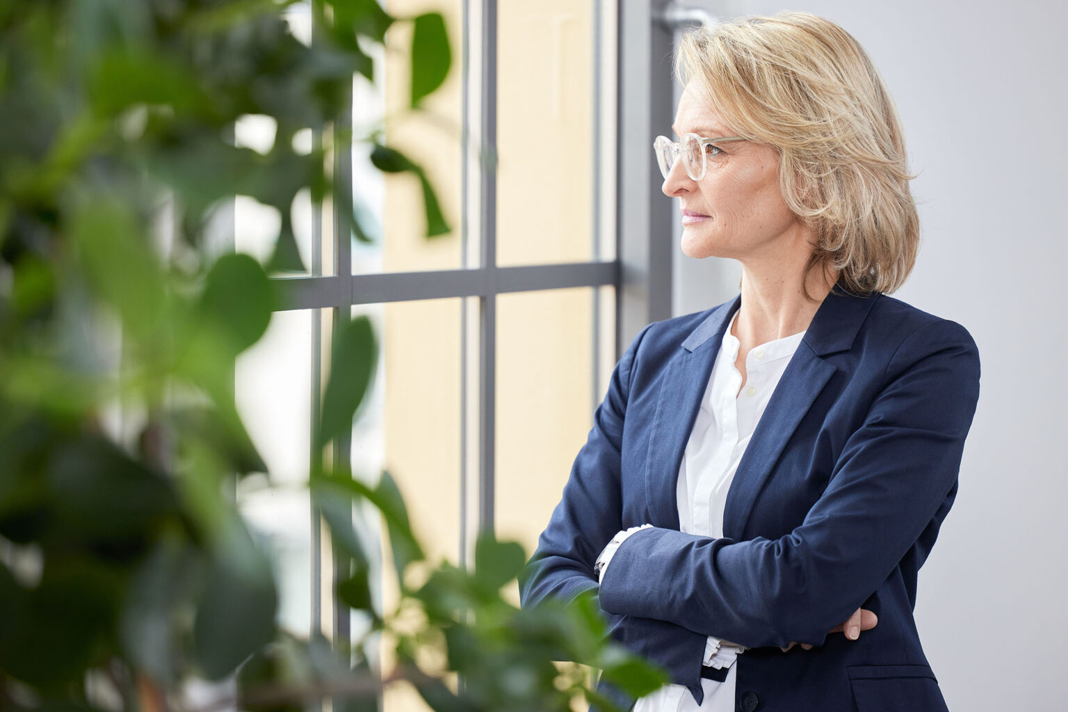 Eine Frau mittleren Alters mit blonden Haaren und Brille steht mit verschränkten Armen vor einem großen Fenster und blickt nach draußen. Sie trägt ein weißes Hemd und ein dunkelblaues Sakko. Links im Bild ist eine grüne Zimmerpflanze zu sehen. Durch das Fenster im Hintergrund sind unscharf Gebäude und die Außenseite zu erkennen.