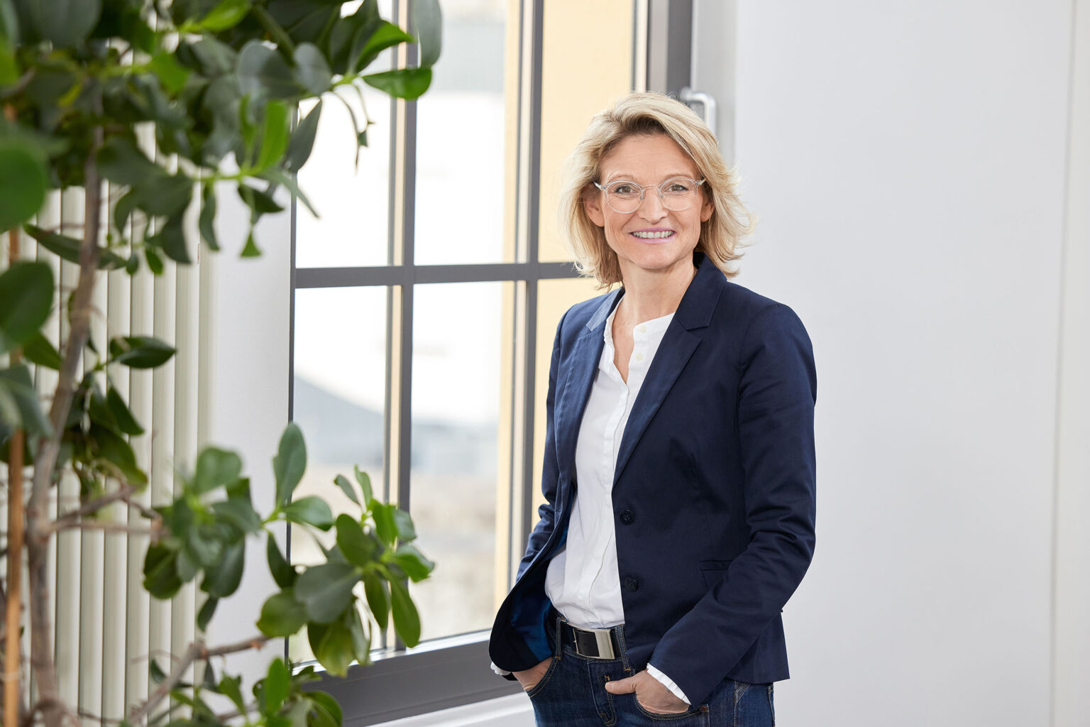 Eine lächelnde Frau mittleren Alters steht vor einem großen Fenster in einem modernen Büro. Sie trägt eine Brille, eine weiße Bluse und ein dunkelblaues Sakko. Ihre Hände sind in die Hosentaschen ihrer dunklen Jeans gesteckt. Links im Bild ist ein Teil einer Zimmerpflanze zu sehen. Der Hintergrund zeigt eine helle, freundliche Büroumgebung.