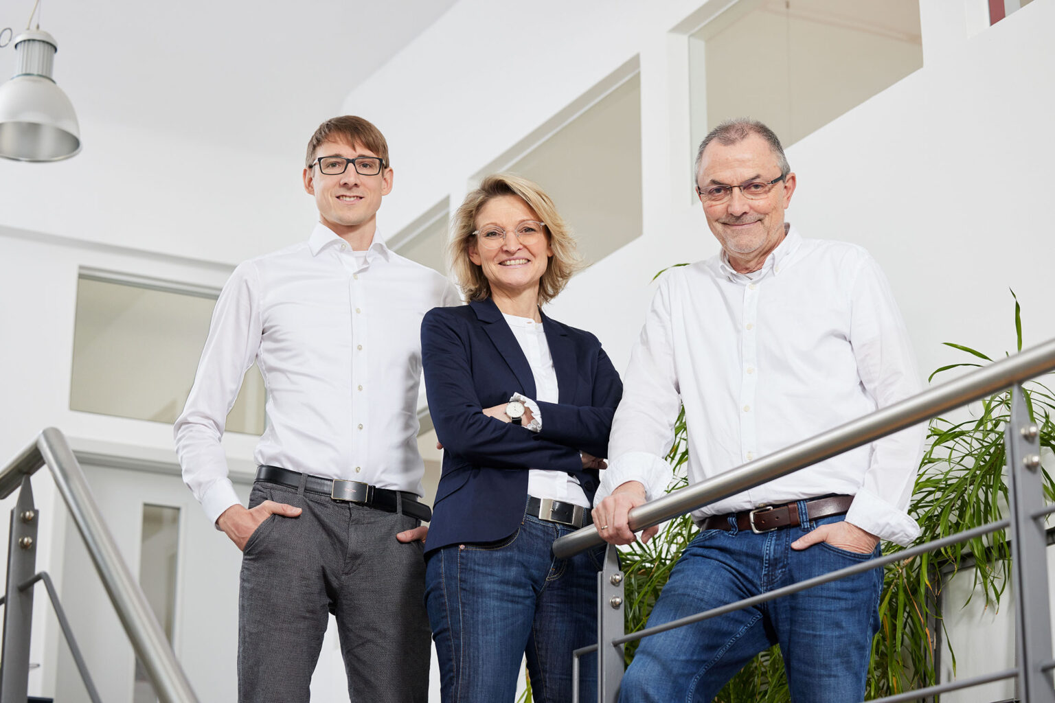 Drei Personen stehen in einem modernen Bürogebäude auf einer Treppe. Links steht ein junger Mann mit Brille, weißem Hemd und grauer Hose, seine Hände sind in den Hosentaschen. In der Mitte steht eine Frau mittleren Alters mit Brille, blondem Haar, dunkelblauem Sakko und weißer Bluse, die ihre Arme verschränkt hat. Rechts steht ein älterer Mann mit Brille, weißem Hemd und blauer Jeans, seine Hände ruhen auf dem Treppengeländer. Im Hintergrund sind weiße Wände und Fenster zu sehen, sowie einige grüne Pflanzen.