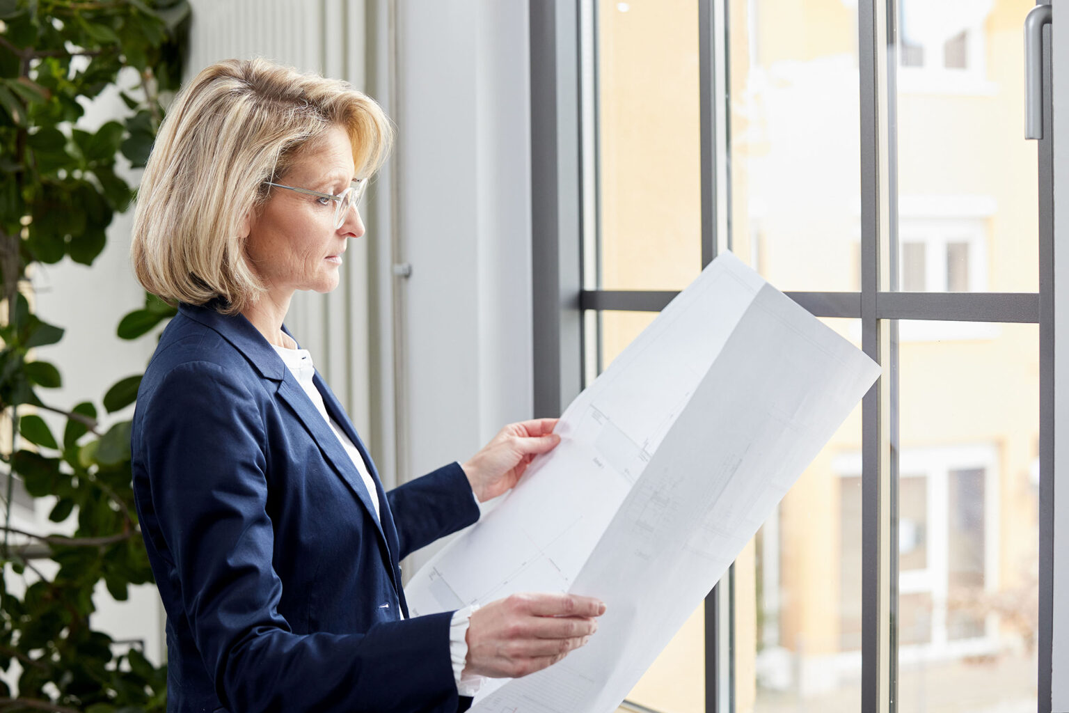 Eine Frau mittleren Alters mit blonden Haaren steht vor einem großen Fenster in einem Büro und betrachtet konzentriert eine große Bauzeichnung. Sie trägt eine Brille, ein weißes Hemd und ein dunkelblaues Sakko. Links im Bild ist eine grüne Zimmerpflanze zu sehen. Durch das Fenster im Hintergrund sind Gebäude und ein Teil der Außenseite zu erkennen.
