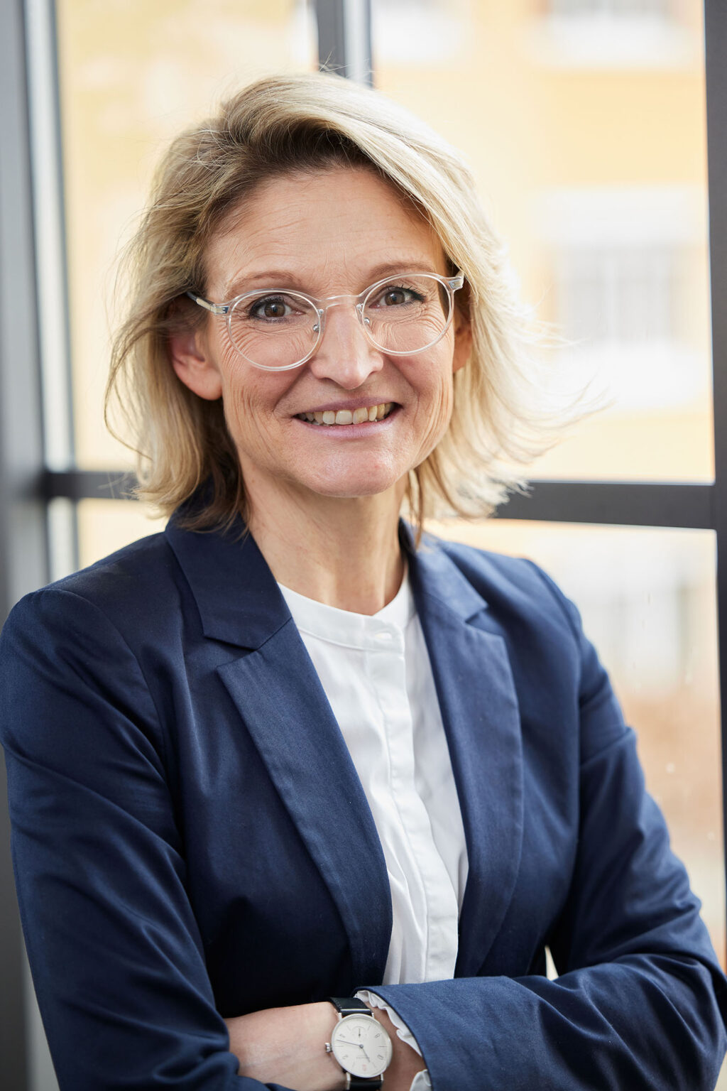 Eine lächelnde Frau mittleren Alters mit blonden Haaren und Brille steht vor einem großen Fenster. Sie trägt ein weißes Hemd und ein dunkelblaues Sakko. Ihre Arme sind vor der Brust verschränkt. Im Hintergrund sind unscharf Gebäude und die Außenseite zu erkennen.