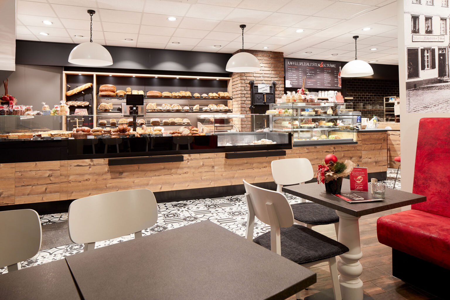 Ein einladender Café- und Bäckereibereich mit einer gemütlichen Atmosphäre. Im Vordergrund befinden sich Tische und Stühle, die zum Verweilen einladen. Ein Tisch ist festlich mit Weihnachtsdekoration geschmückt. Im Hintergrund ist eine große Verkaufstheke aus Holz zu sehen, die mit verschiedenen Backwaren wie Brot, Brötchen und Gebäck gefüllt ist. Über der Theke hängen weiße Pendelleuchten, die den Bereich gut beleuchten. Die Wände sind teilweise mit Ziegelsteinen verkleidet und es gibt dekorative Schwarz-Weiß-Fotos. Der Boden ist mit dekorativen Fliesen gestaltet.