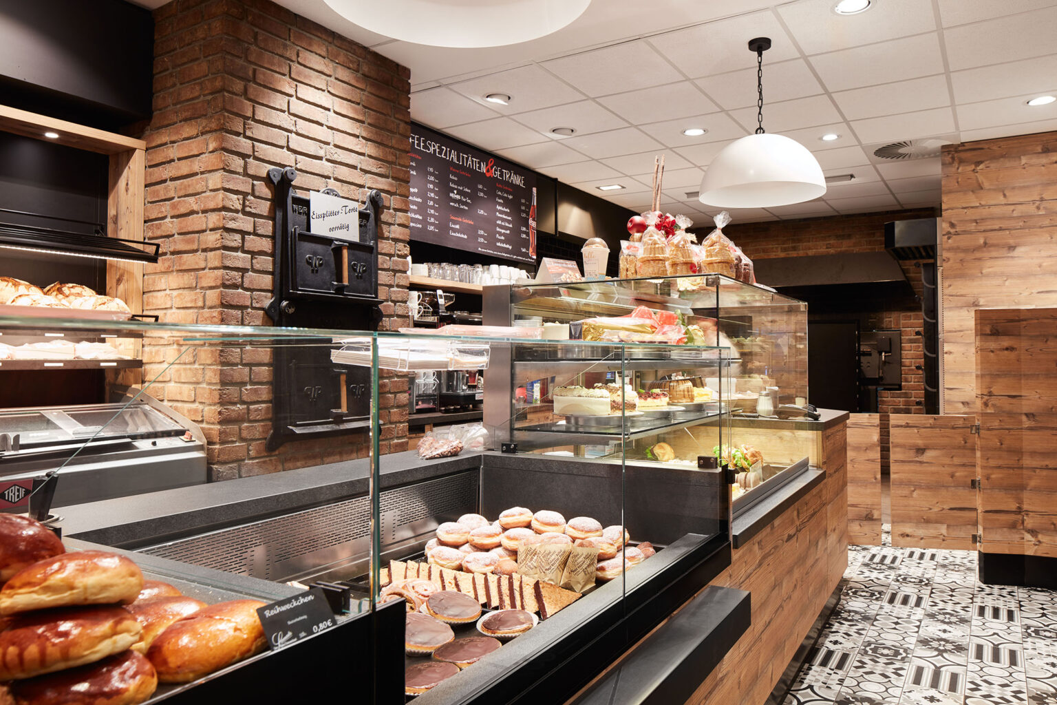 Ein gemütlicher Bäckereibereich mit einer rustikalen und einladenden Atmosphäre. Die Wände sind mit Ziegelsteinen verkleidet, und die Einrichtung besteht aus Holz. Hinter der Theke befinden sich Glasvitrinen, die eine Vielzahl von Backwaren und Desserts präsentieren, darunter Brötchen, Kuchen und Gebäck. Über der Theke hängen Menüs, die verschiedene Spezialitäten und Getränke anzeigen. Der Boden ist mit dekorativen Fliesen ausgelegt. Die Beleuchtung ist warm und trägt zur einladenden Atmosphäre des Raumes bei.