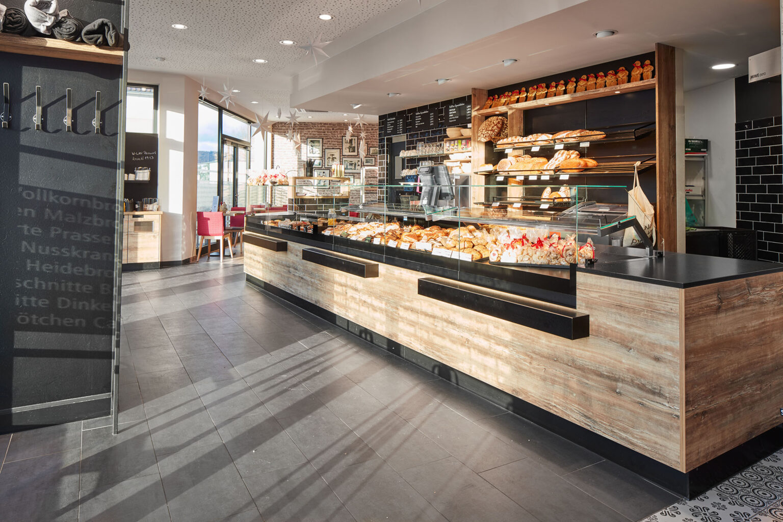 Ein heller und einladender Bäckereibereich mit großen Glasvitrinen, die eine Vielzahl von Backwaren wie Brot, Brötchen und Gebäck präsentieren. Die Theke und Regale sind aus hellem Holz gefertigt, was eine warme und rustikale Atmosphäre schafft. Im Hintergrund sind Wände mit Ziegelverkleidung und dekorativen Elementen zu sehen. Links im Bild befindet sich ein Sitzbereich mit roten Polsterstühlen und Tischen. Der Boden ist mit dunklen Fliesen ausgelegt und der Raum ist gut beleuchtet, mit viel natürlichem Licht, das durch große Fenster hereinfällt.