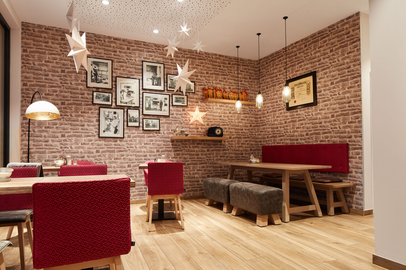 Ein gemütlicher Essbereich in einem Restaurant mit einer warmen und einladenden Atmosphäre. Die Wände sind mit Ziegelsteinen verkleidet und mit gerahmten Schwarz-Weiß-Fotografien sowie dekorativen Sternen geschmückt. Der Raum ist mit Holzmöbeln ausgestattet, darunter Tische, rote Polsterstühle und Holzbänke mit gepolsterten Hockern. Über den Tischen hängen verschiedene Lampen, die den Raum in warmes Licht tauchen. Eine hölzerne Regalfläche an der Wand zeigt dekorative Objekte und eine Vintage-Radio. Der Boden ist aus hellem Holz, was zur gemütlichen Atmosphäre beiträgt.