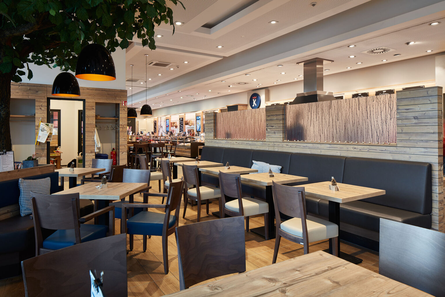 Ein geräumiger und gemütlicher Restaurantbereich mit Holztischen und Stühlen, die mit blauen Polstern ausgestattet sind. An den Wänden sind dekorative Elemente und Trennwände aus Holz zu sehen. Im Vordergrund stehen mehrere Tische, die für Gäste vorbereitet sind. Über den Tischen hängen moderne schwarze Pendelleuchten mit warmem Licht. Ein großer Baum mit grünen Blättern steht links im Bild und trägt zur gemütlichen Atmosphäre bei. Im Hintergrund sind weitere Sitzbereiche und eine Theke zu sehen