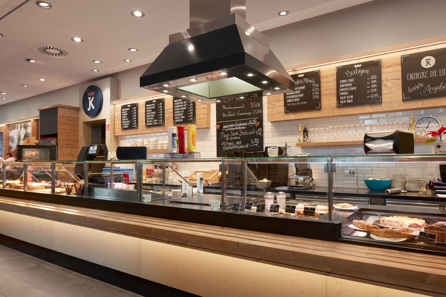 Ein gut beleuchteter Verkaufstresen in einem modernen Restaurant oder Café. Der Tresen ist mit Glasvitrinen ausgestattet, die verschiedene Backwaren, Kuchen und Desserts präsentieren. Über dem Tresen hängen Menüs auf Holztafeln, die verschiedene Speisen und Getränke anzeigen. Eine Dunstabzugshaube hängt über dem Bereich, wo Speisen zubereitet werden. Die Wände sind mit weißen Fliesen verkleidet, und es gibt Regale mit Flaschen, Gläsern und anderen Utensilien. Die Einrichtung ist eine Kombination aus Holz und modernen Elementen, was eine einladende Atmosphäre schafft.