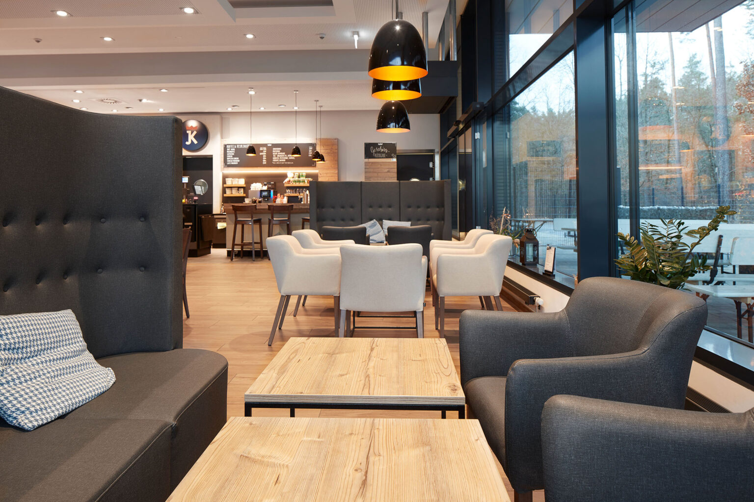 Ein gemütlicher Sitzbereich in einem modernen Café oder Restaurant. Der Bereich ist mit grauen und weißen Polstersesseln sowie hellen Holztischen ausgestattet. Große Fenster lassen viel natürliches Licht in den Raum und bieten einen Blick auf die Außenterrasse. Über den Tischen hängen schwarze Pendelleuchten mit warmem Licht. Im Hintergrund sind weitere Sitzbereiche und eine Theke zu sehen. Die Einrichtung ist modern und einladend, mit einem Mix aus dunklen und hellen Farbtönen
