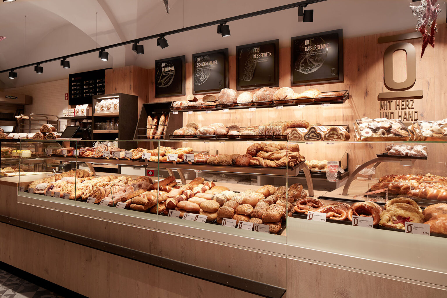 Ein gut beleuchteter Bäckereibereich mit einer Vielzahl von Backwaren, die in Glasvitrinen ausgestellt sind. Die Auslage zeigt eine große Auswahl an Brot, Brötchen, Brezeln und Gebäck. Über den Vitrinen sind Holzregale mit weiteren Backwaren und dekorativen Schildern angebracht. An der Wand hinter der Theke befindet sich das Logo der Bäckerei mit dem Slogan 'Mit Herz und Hand'. Der Boden ist mit dekorativen Fliesen gestaltet und die Decke ist in einem eleganten Bogen gehalten. Die Atmosphäre ist warm und einladend.