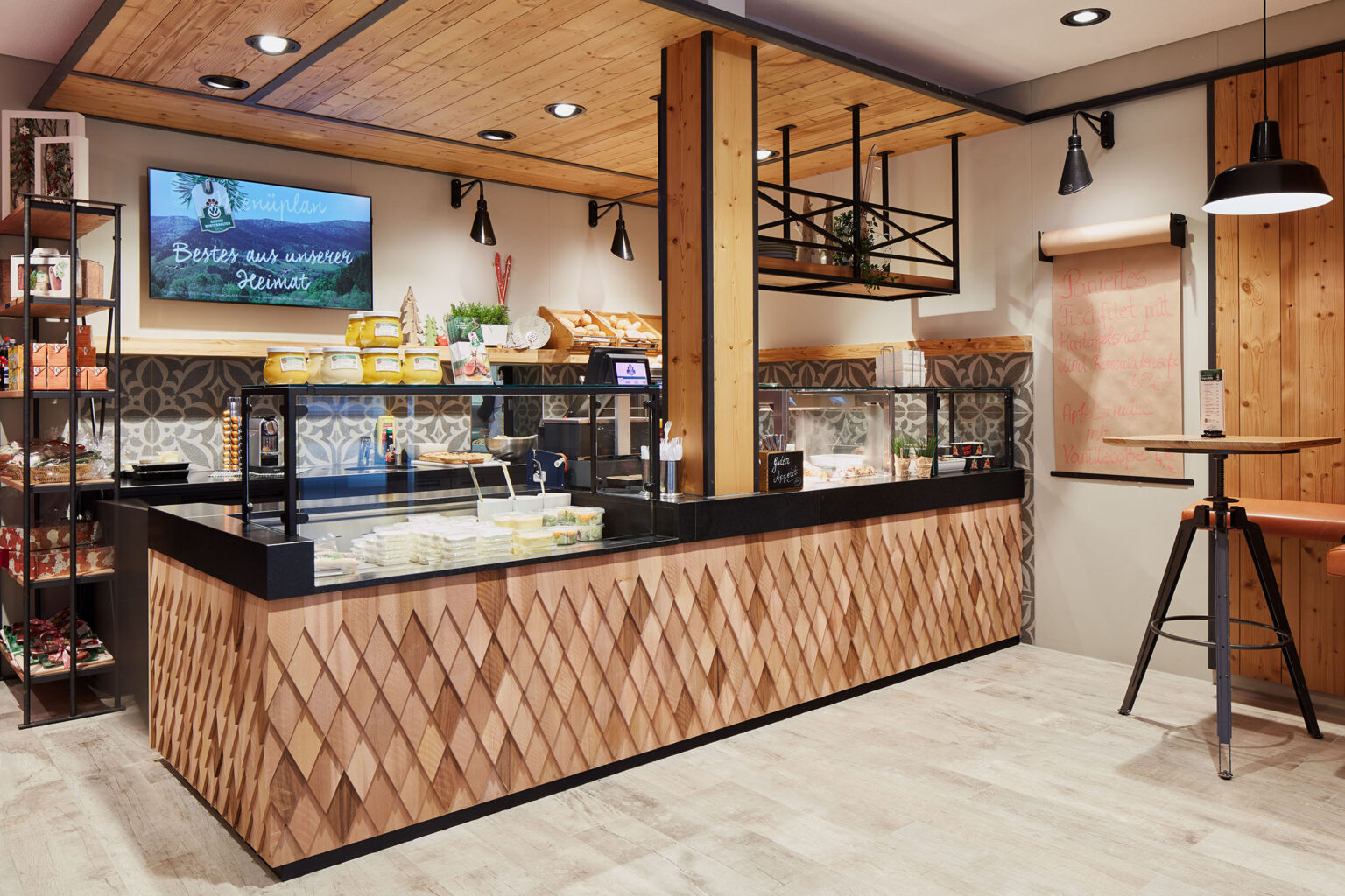 Ein gemütlicher und rustikaler Verkaufsbereich in einem Geschäft, dekoriert mit Holzelementen und modernen Akzenten. Im Vordergrund befindet sich eine Theke mit einer kunstvollen Holzverkleidung und einer Glasvitrine, die verschiedene frische Lebensmittel präsentiert. Im Hintergrund sind Regale mit Backwaren und weiteren Produkten zu sehen. An der Wand hängt ein Bildschirm mit der Aufschrift 'Bester aus unserer Heimat'. Die Decke ist ebenfalls mit Holz verkleidet und an der Wand hängt eine Rolle mit einem handgeschriebenen Menü. Der Boden ist aus hellem Holz, was die warme Atmosphäre des Raumes verstärkt.