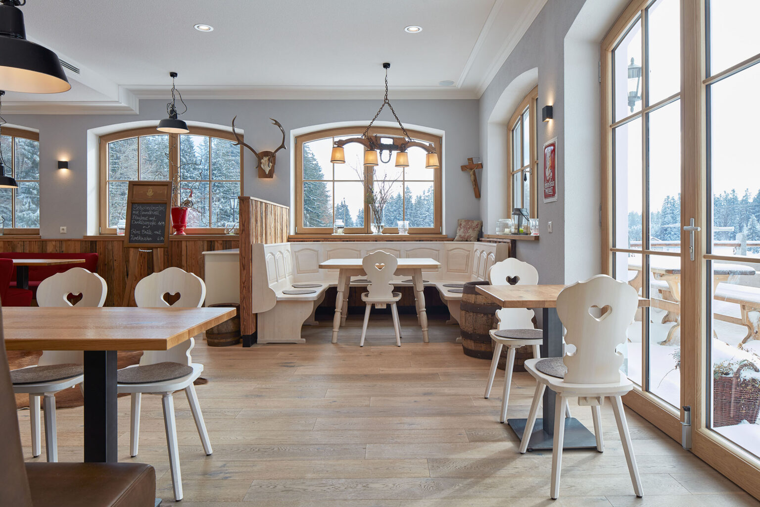 Ein gemütliches Restaurant mit großen Fenstern, durch die eine schneebedeckte Landschaft zu sehen ist. Der Raum ist mit Holztischen und weißen Stühlen mit herzförmigen Rückenlehnen ausgestattet. In einer Ecke befindet sich eine rustikale Sitzbank mit passendem Tisch. An den Wänden hängen Dekorationen wie ein Hirschgeweih, eine hölzerne Kreuz-Dekoration und ein Schild mit Menüangeboten. Die warme, einladende Atmosphäre wird durch Holzvertäfelungen und weiche Polster betont.