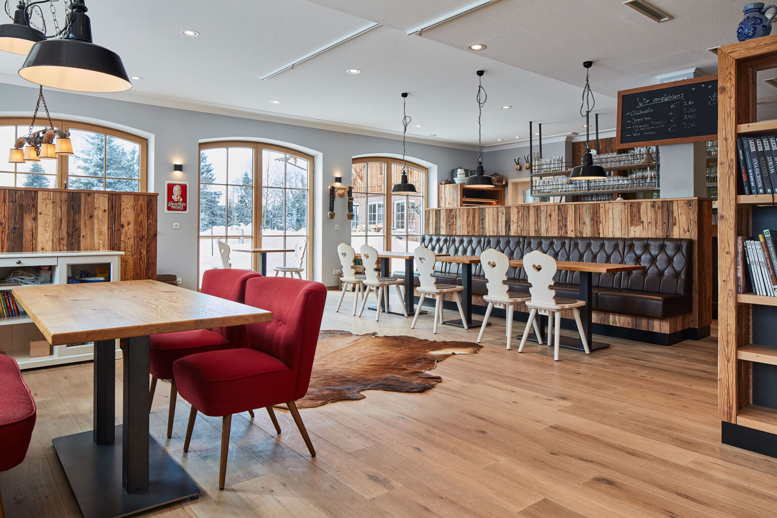Ein gemütlich eingerichtetes Restaurant mit großen Fenstern, durch die eine schneebedeckte Landschaft zu sehen ist. Der Raum ist mit Holztischen, roten Polstersesseln und weißen Stühlen mit herzförmigen Rückenlehnen ausgestattet. An den Wänden hängen rustikale Dekorationen, wie Kuhglocken und ein Schild mit Menüangeboten. Eine Theke mit Barhockern und hängenden Lampen sorgt für eine einladende Atmosphäre. Ein Regal mit Büchern und eine weiße Kommode mit Spielzeug und Büchern sind ebenfalls im Raum.