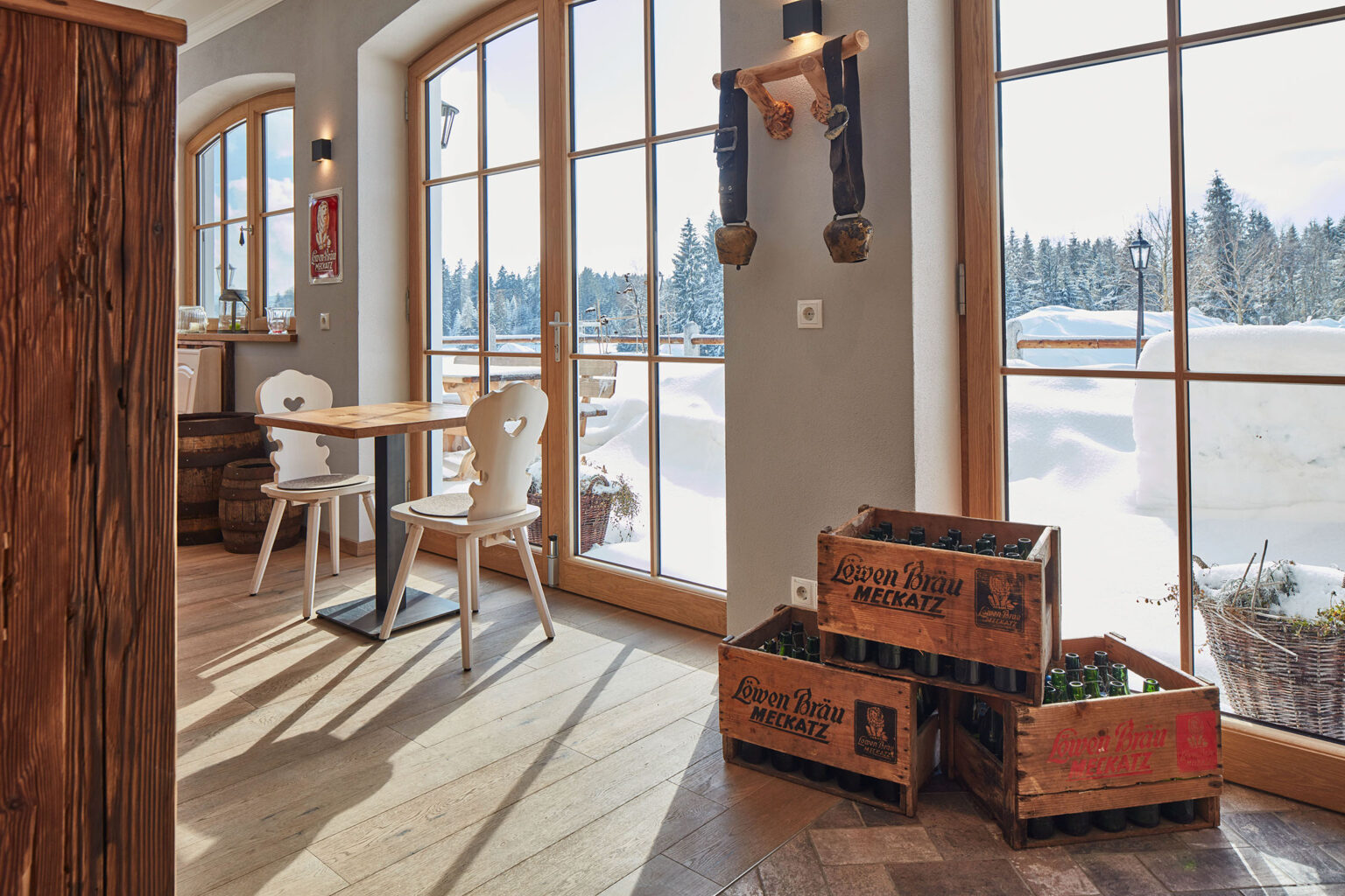 Ein heller Raum mit großen Fenstern, die einen Blick auf eine schneebedeckte Landschaft bieten. Im Raum stehen weiße Holzstühle und Holztische auf einem Holzboden. An der Wand hängt ein Schild mit der Aufschrift 'Löwenbräu Meckatz' sowie eine traditionelle Glocke. Auf dem Boden stehen mehrere Holzkisten mit leeren Bierflaschen der Marke 'Löwenbräu Meckatz'. Der Raum ist gemütlich und rustikal eingerichtet.